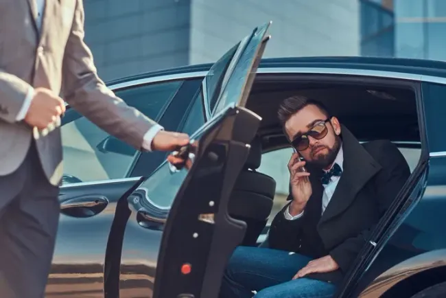 Man in sunglasses talking on phone while seated in a black car as a driver opens the door