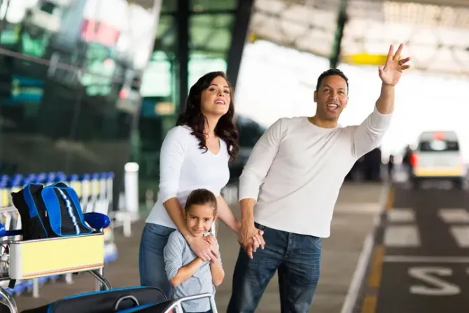 Family waving at an airport curb with luggage carts and cars in the background