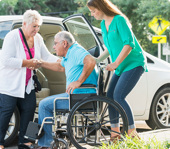 Two caregivers assisting a wheelchair user beside an open car trunk outdoors