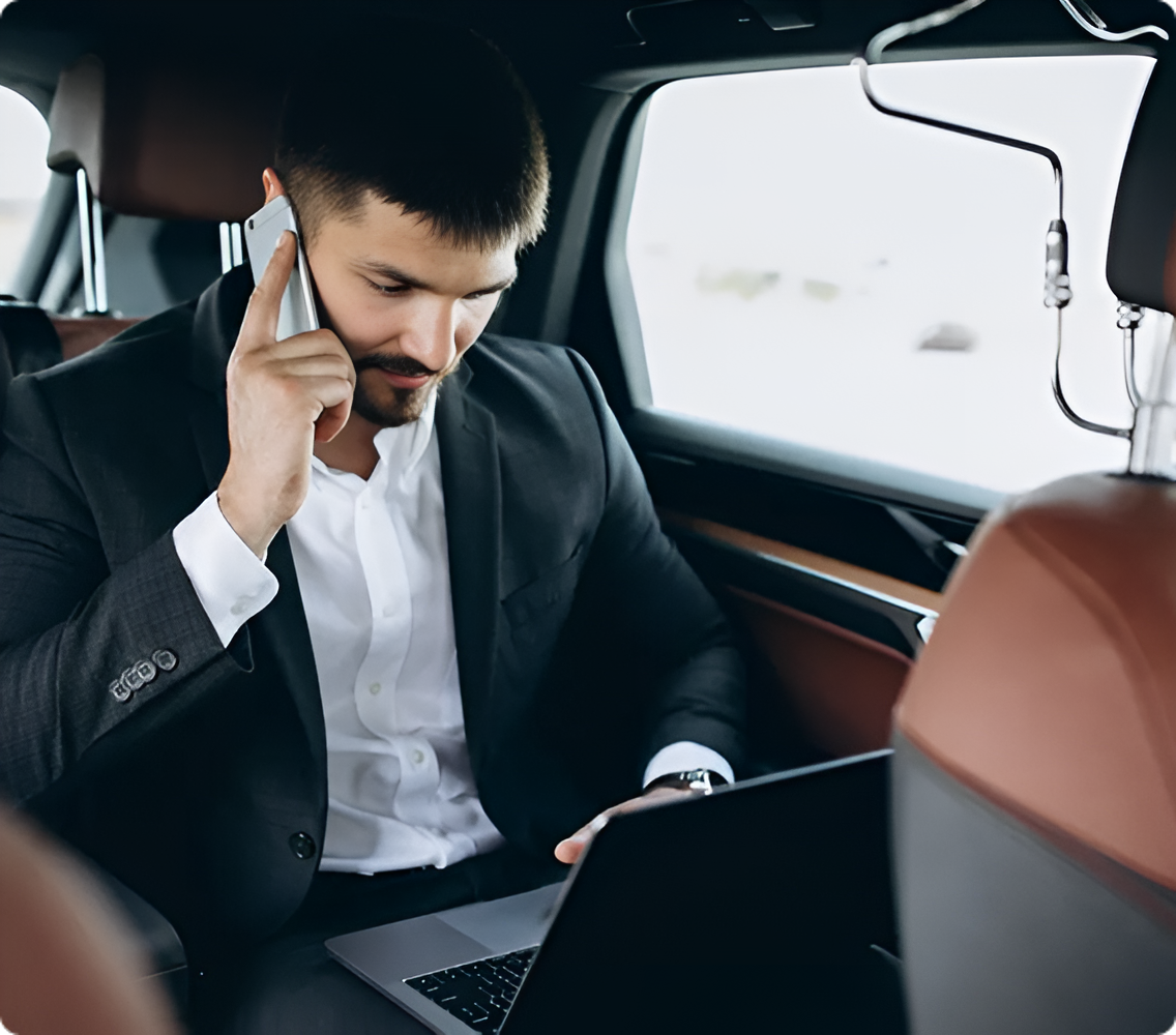 Man in a black suit using a laptop and talking on a phone inside a car
