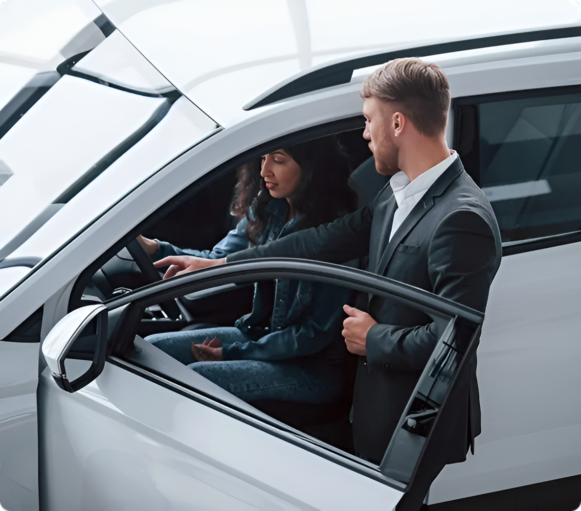 Two people inside a white car, one leaning in through the open driver’s door and pointing at the dashboard.