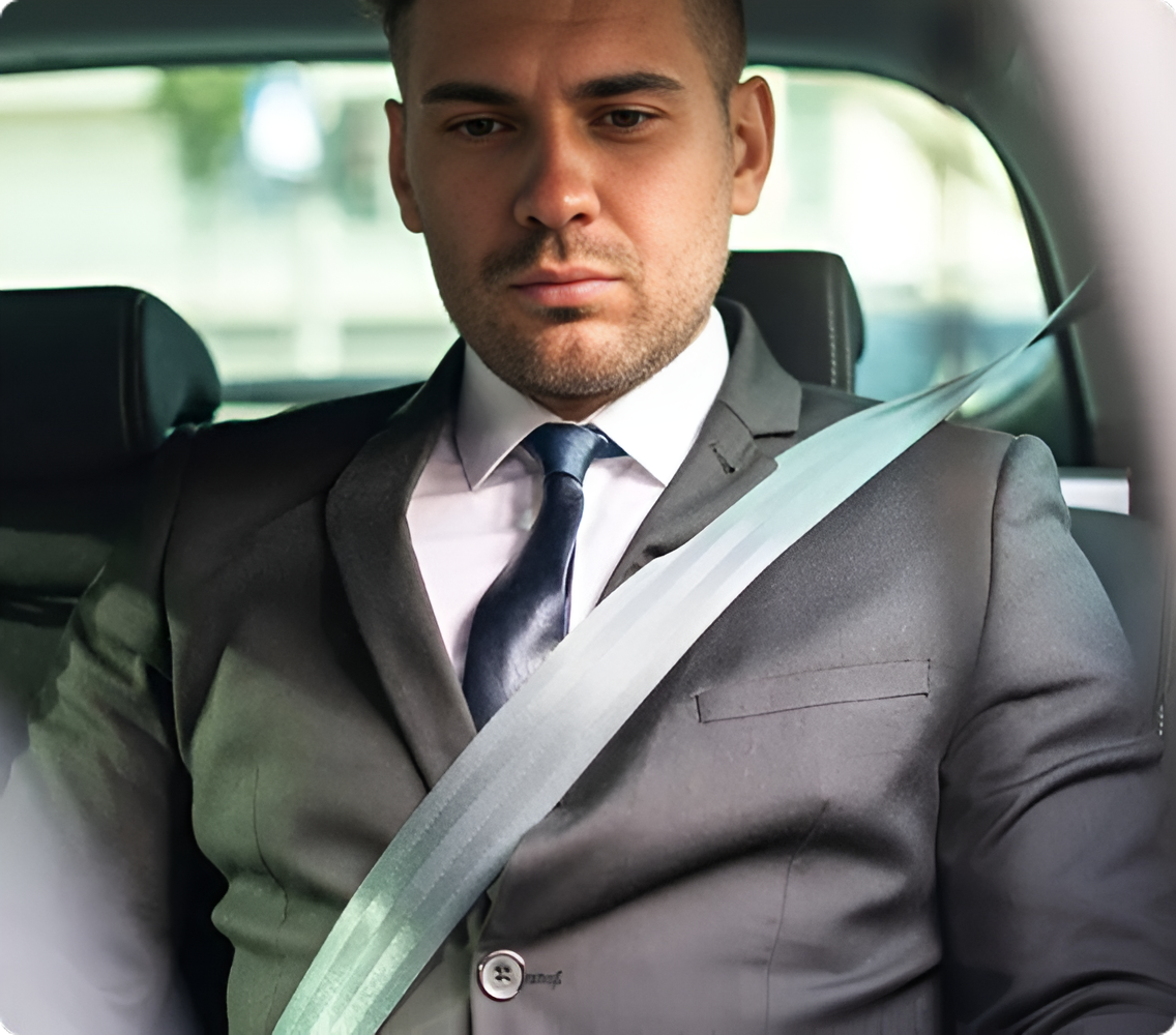 Man in a gray suit sitting in a car, looking serious with a seat belt on.