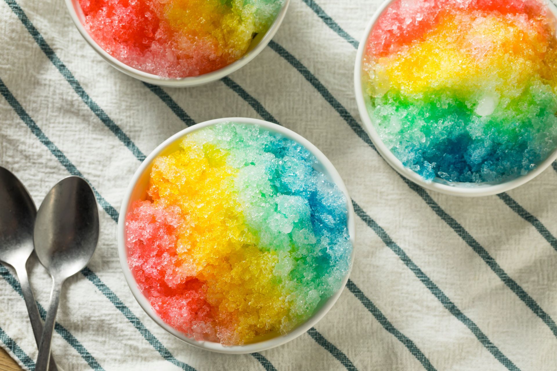 Three bowls of colorful shaved ice with spoons on a table.