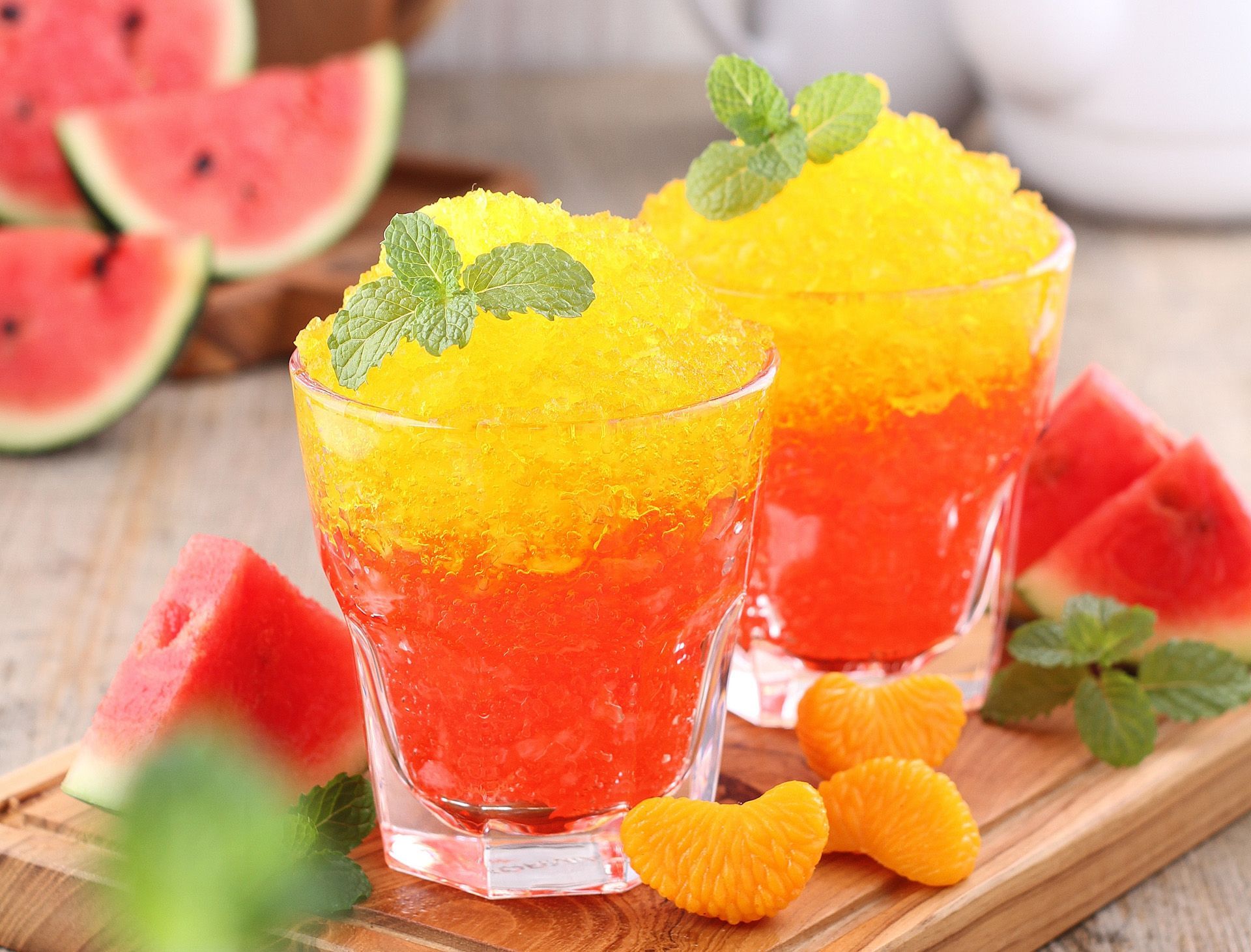 Two glasses of slush with watermelon and orange slices on a wooden cutting board.