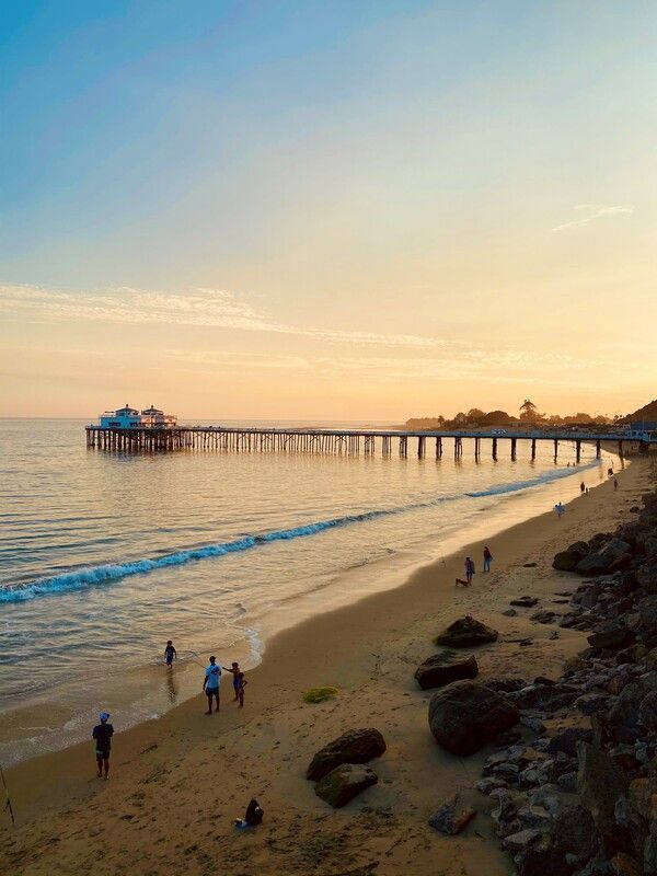 A group of people are standing on a beach near a pier at sunset.