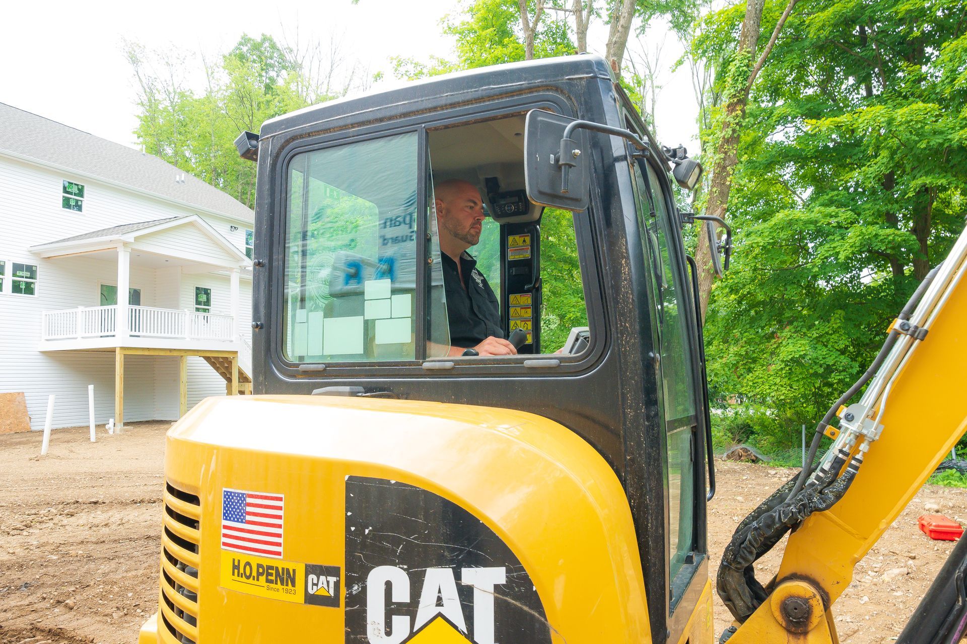 Yellow CAT excavator with a driver inside, parked beside a house and trees.