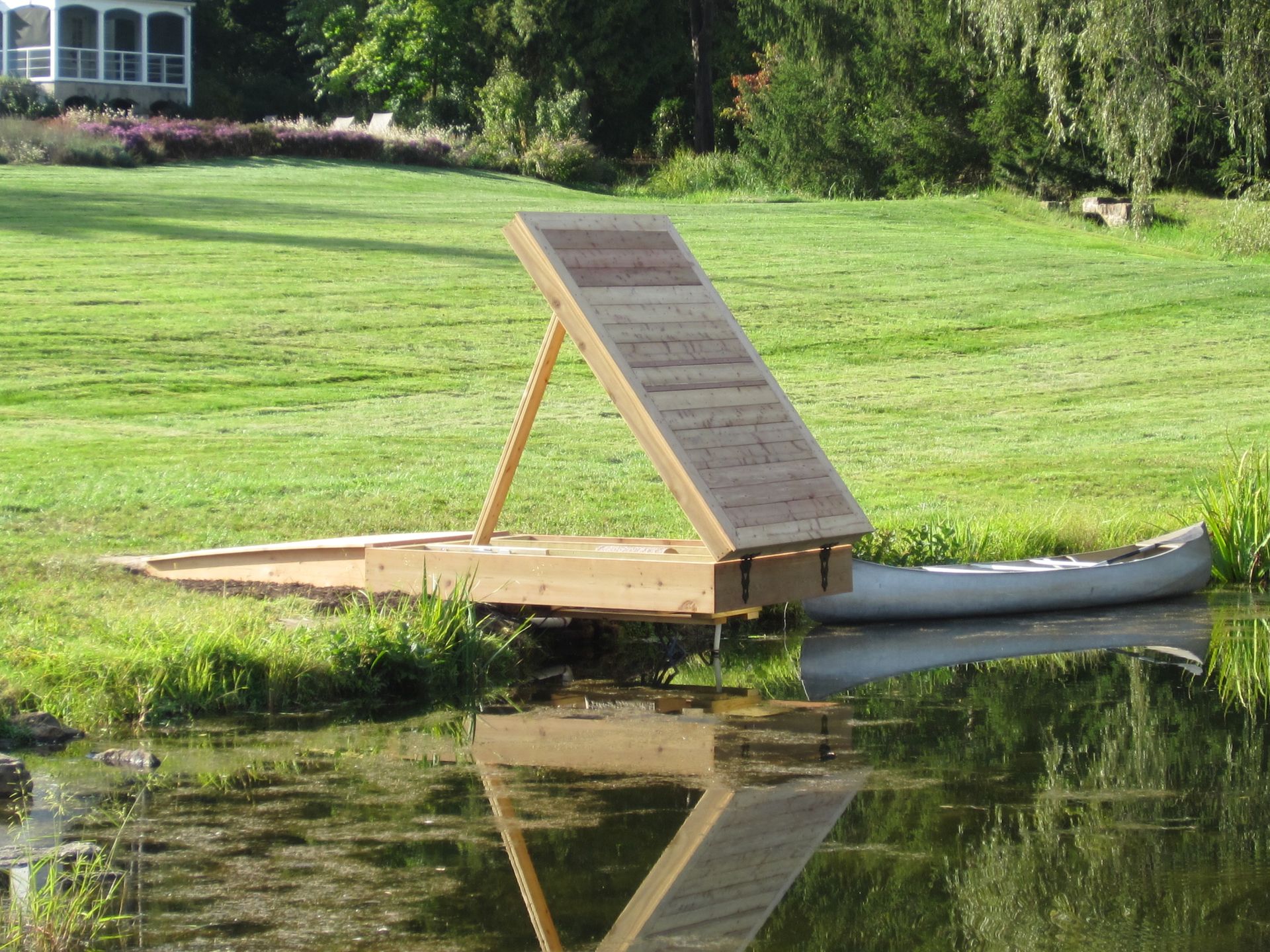 Floating wooden ramp on a pond beside a grassy shore, with a small canoe and tree reflections.