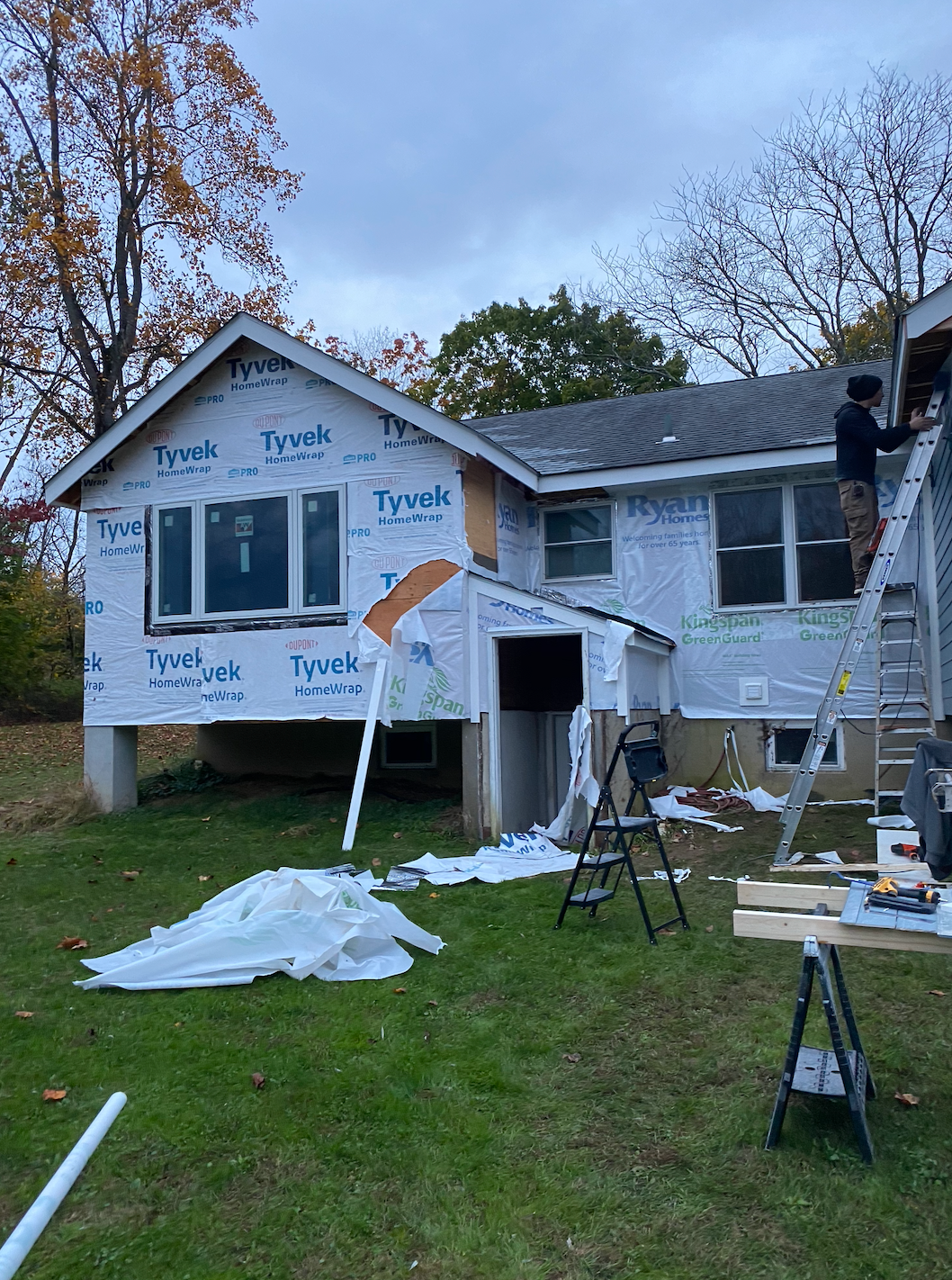Workers install siding on a two-story house wrapped in Tyvek, with ladders and tools in the yard.