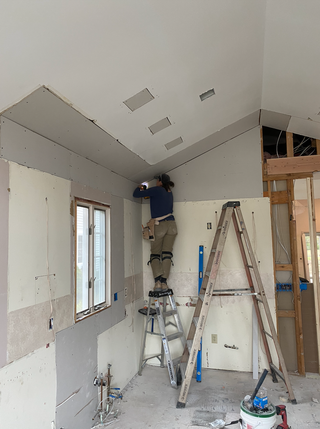 Renovation room with drywall, ladders, tools, and a worker standing on a ladder near the ceiling