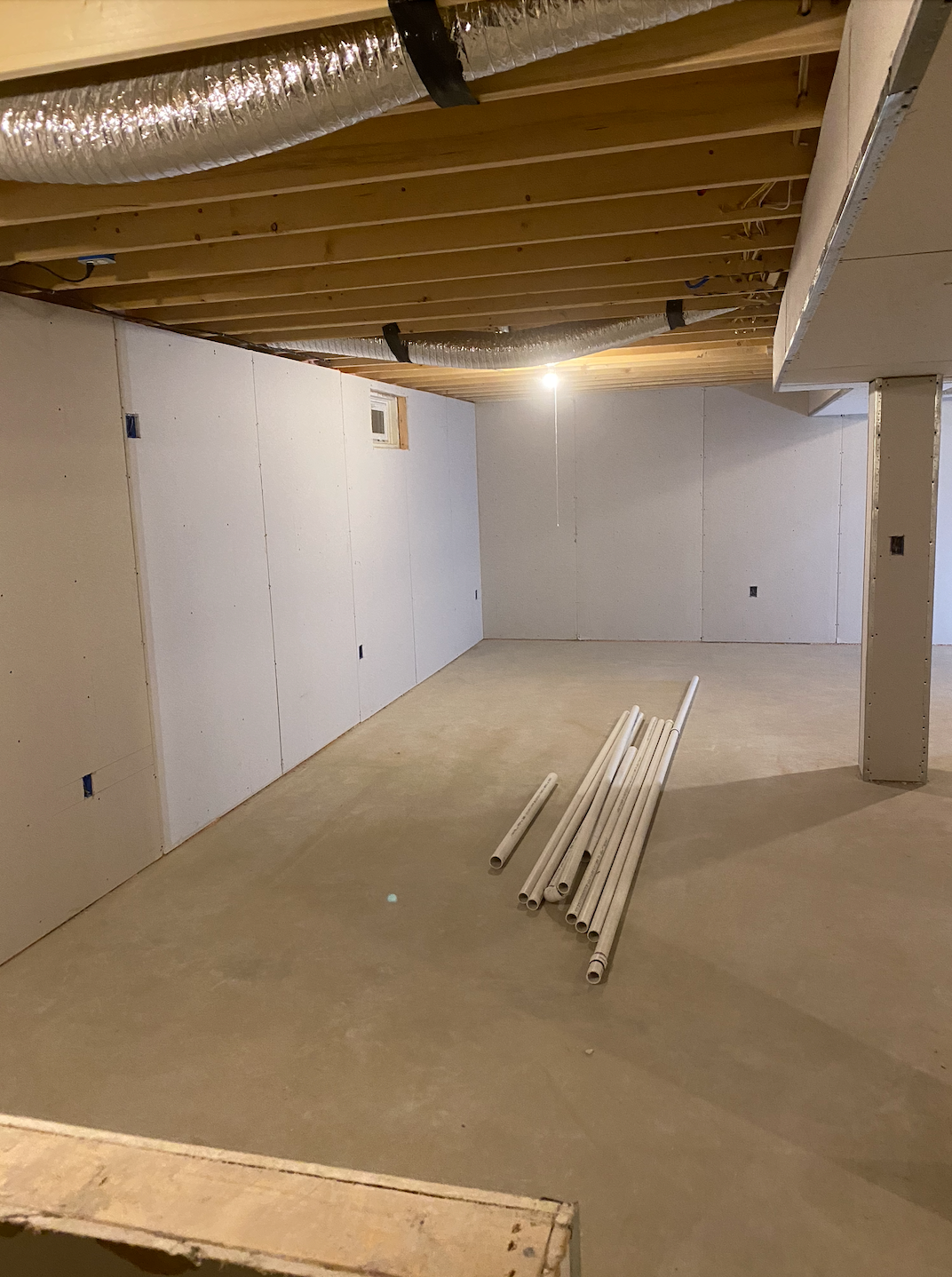 Empty unfinished basement with white walls, exposed ceiling, concrete floor, and stacked white trim pieces.