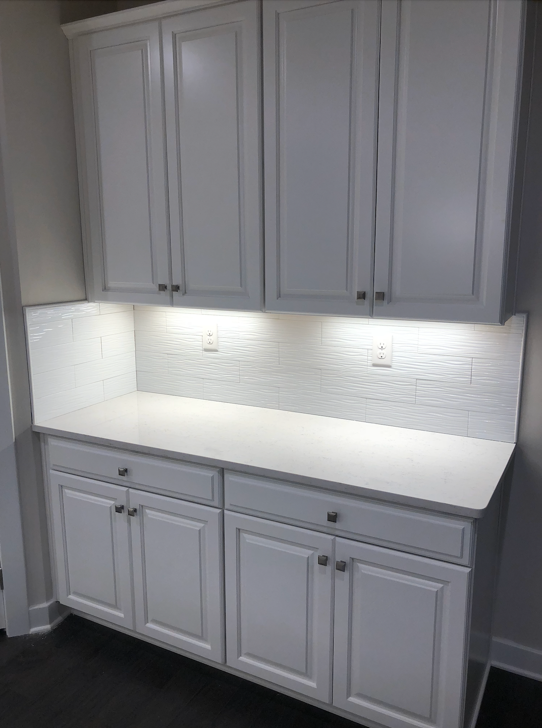 White built-in cabinets and counter with under-cabinet lighting against a textured white backsplash.
