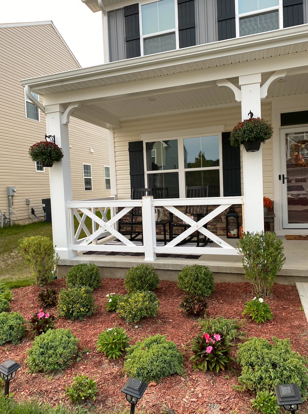 Front porch with white railings, hanging flower baskets, and a landscaped garden in front of a beige house