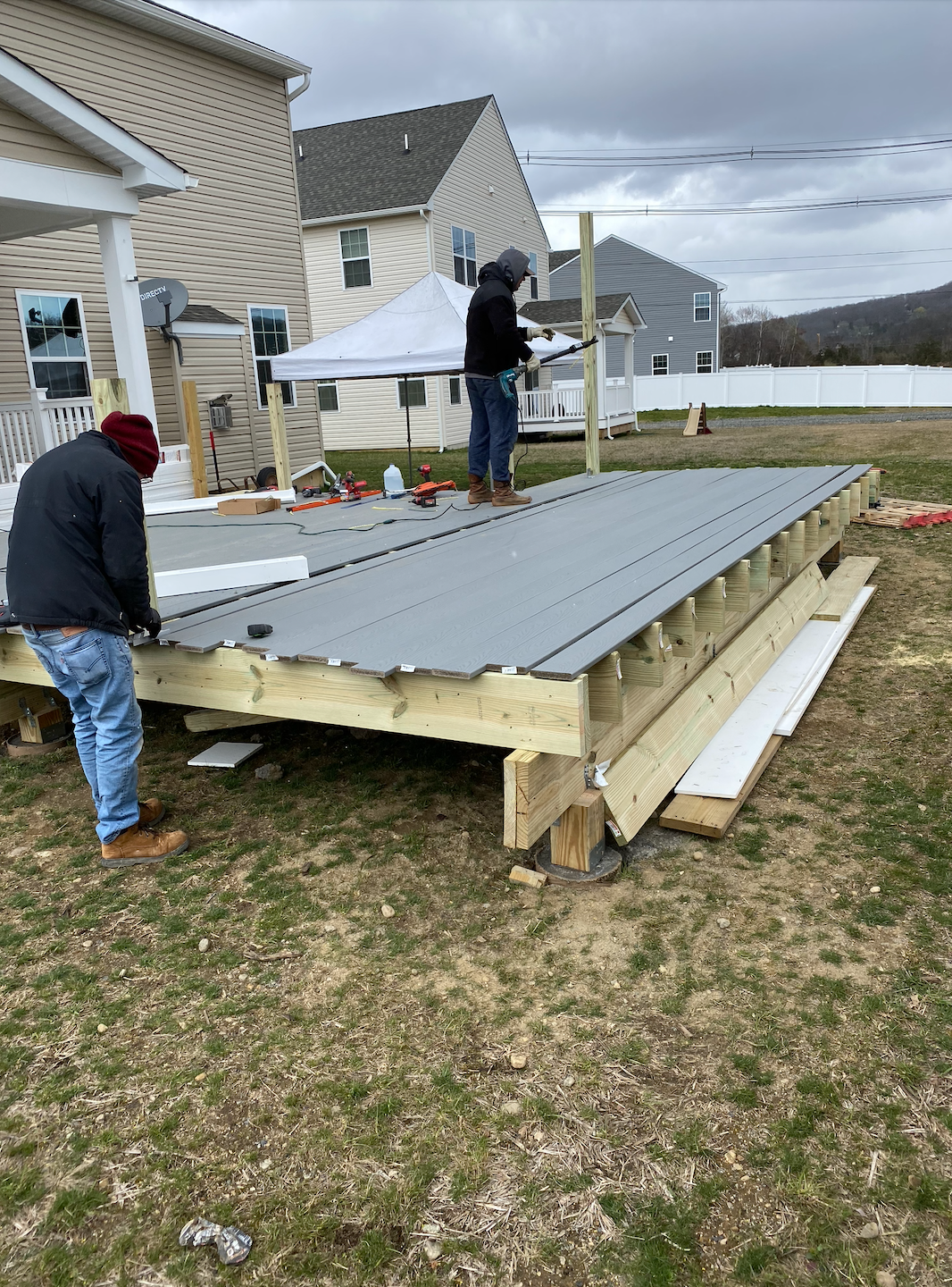 Workers installing gray metal roof panels on a raised deck beside a house, with a tent and neighborhood in the background
