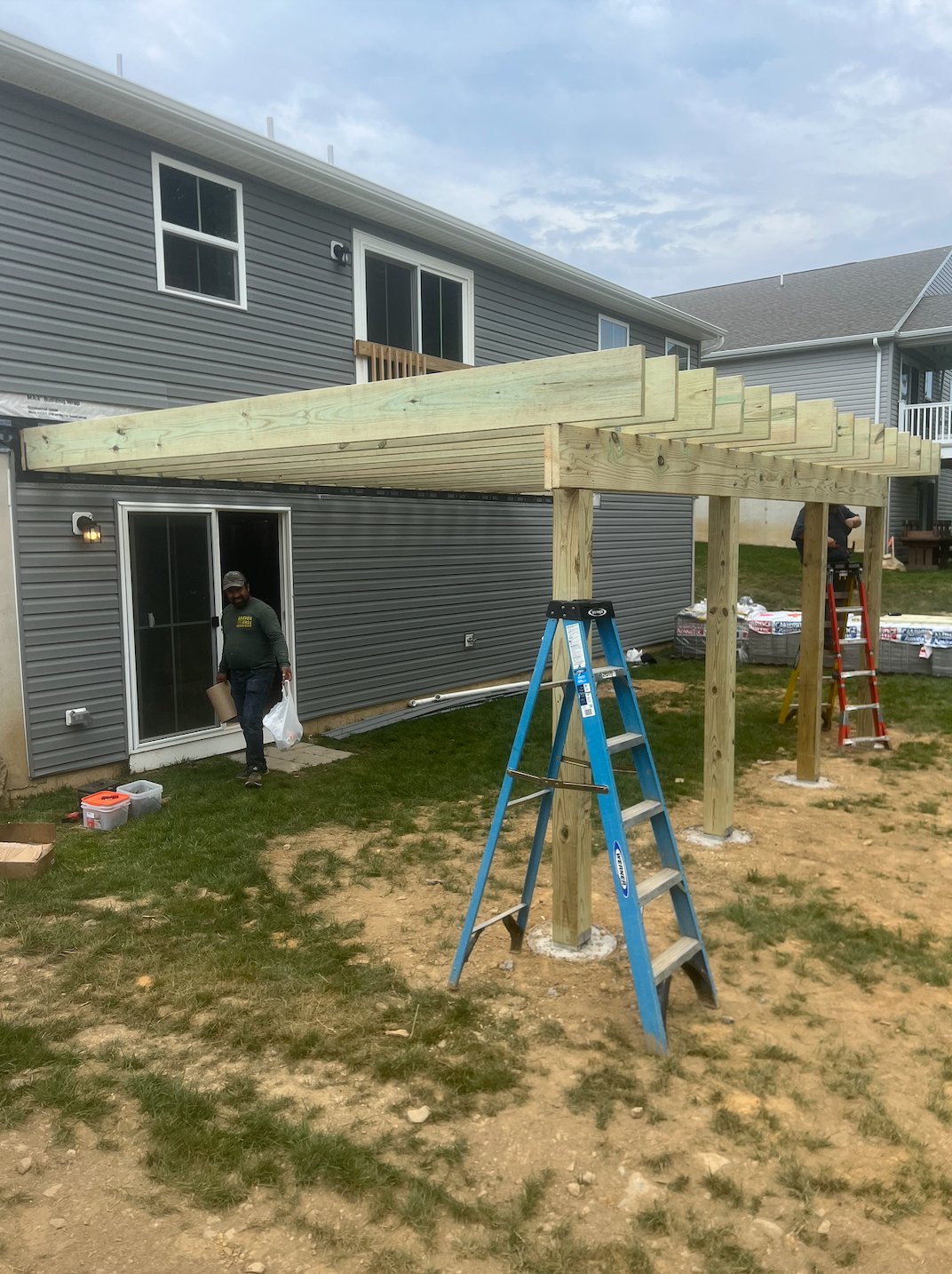 Backyard deck framing under construction beside a gray house, with tools and ladders nearby.