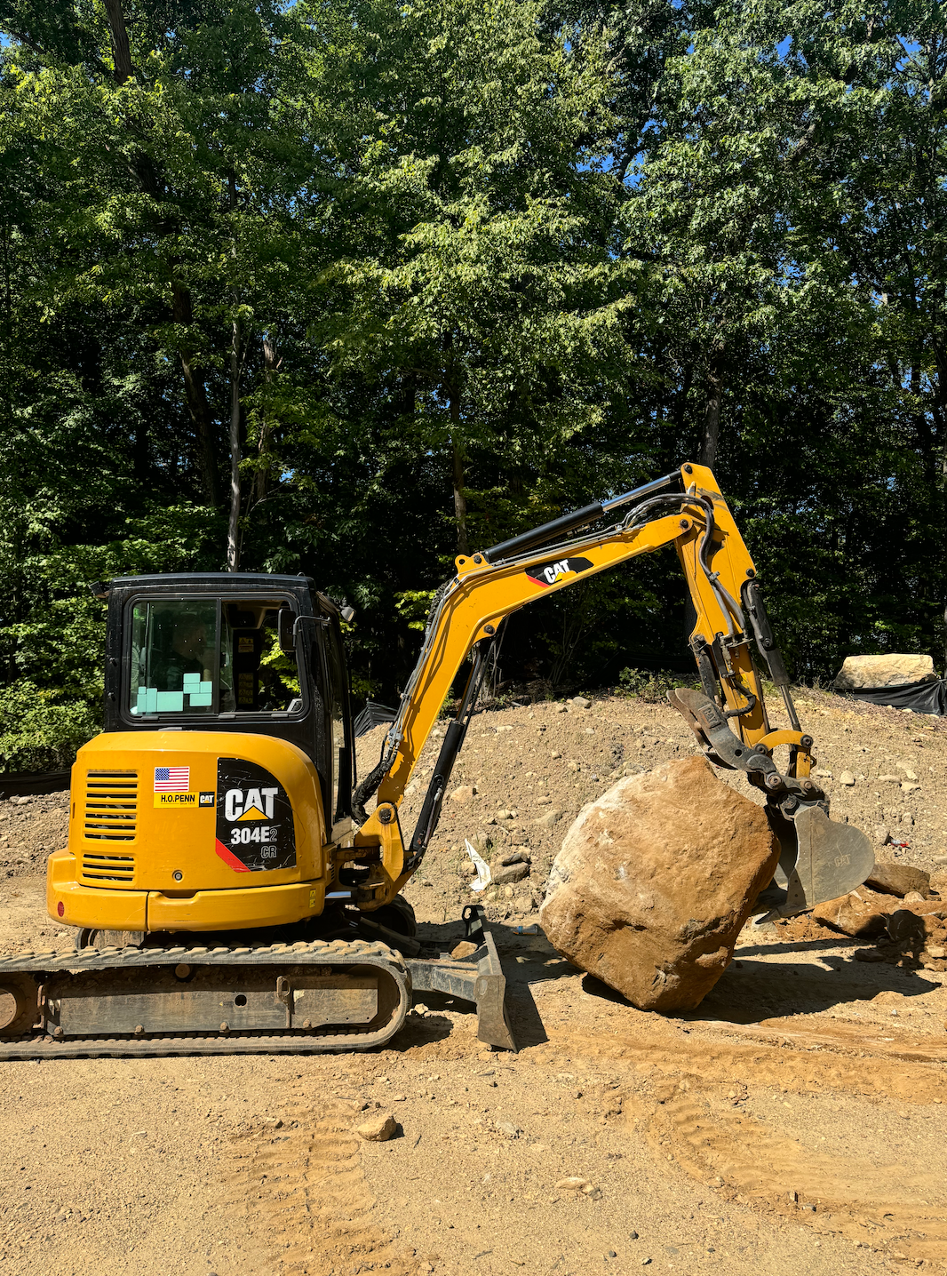 Yellow excavator lifting a large boulder on a dirt site with trees in the background