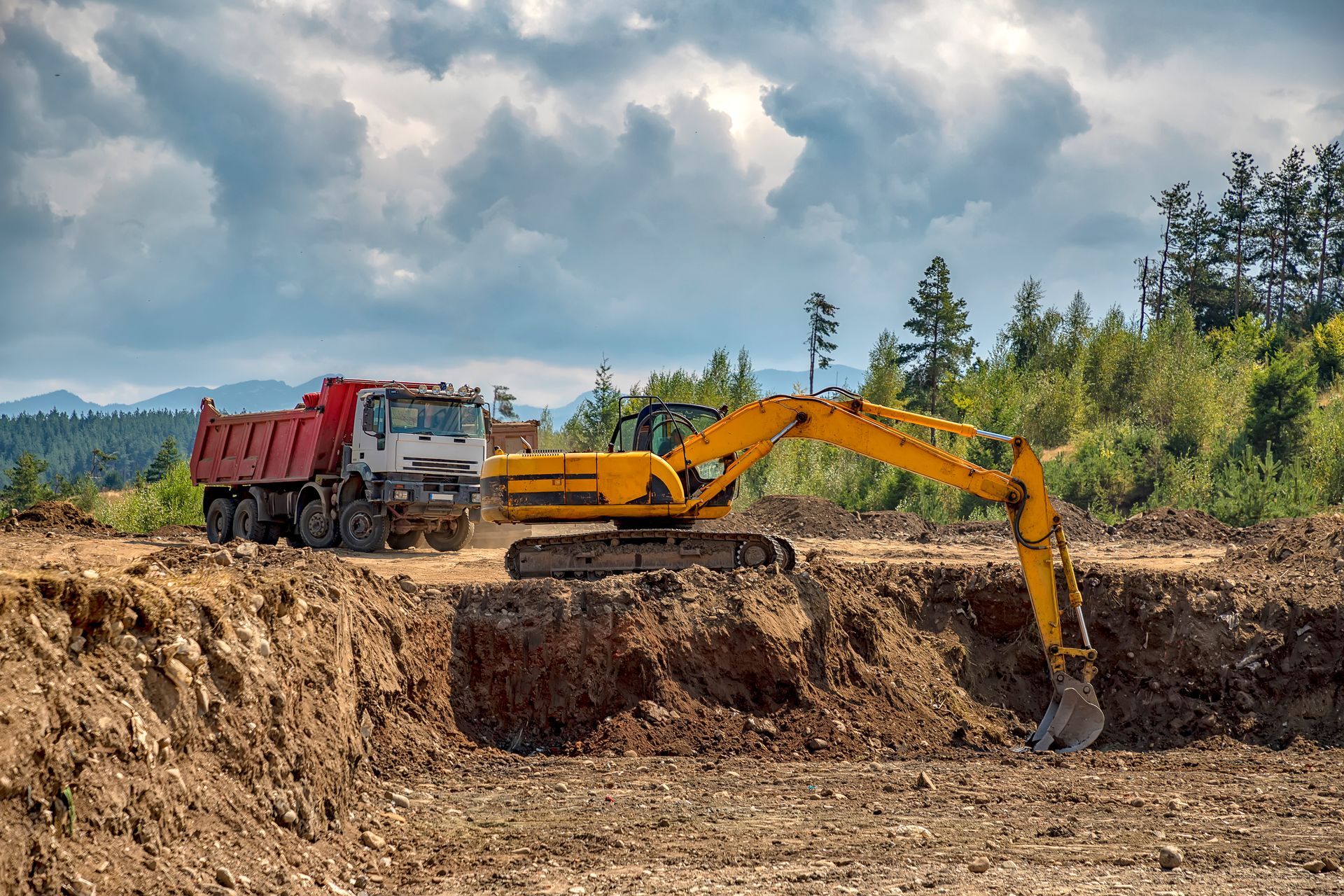 Yellow excavator digging earth beside a red dump truck at a construction site under cloudy skies