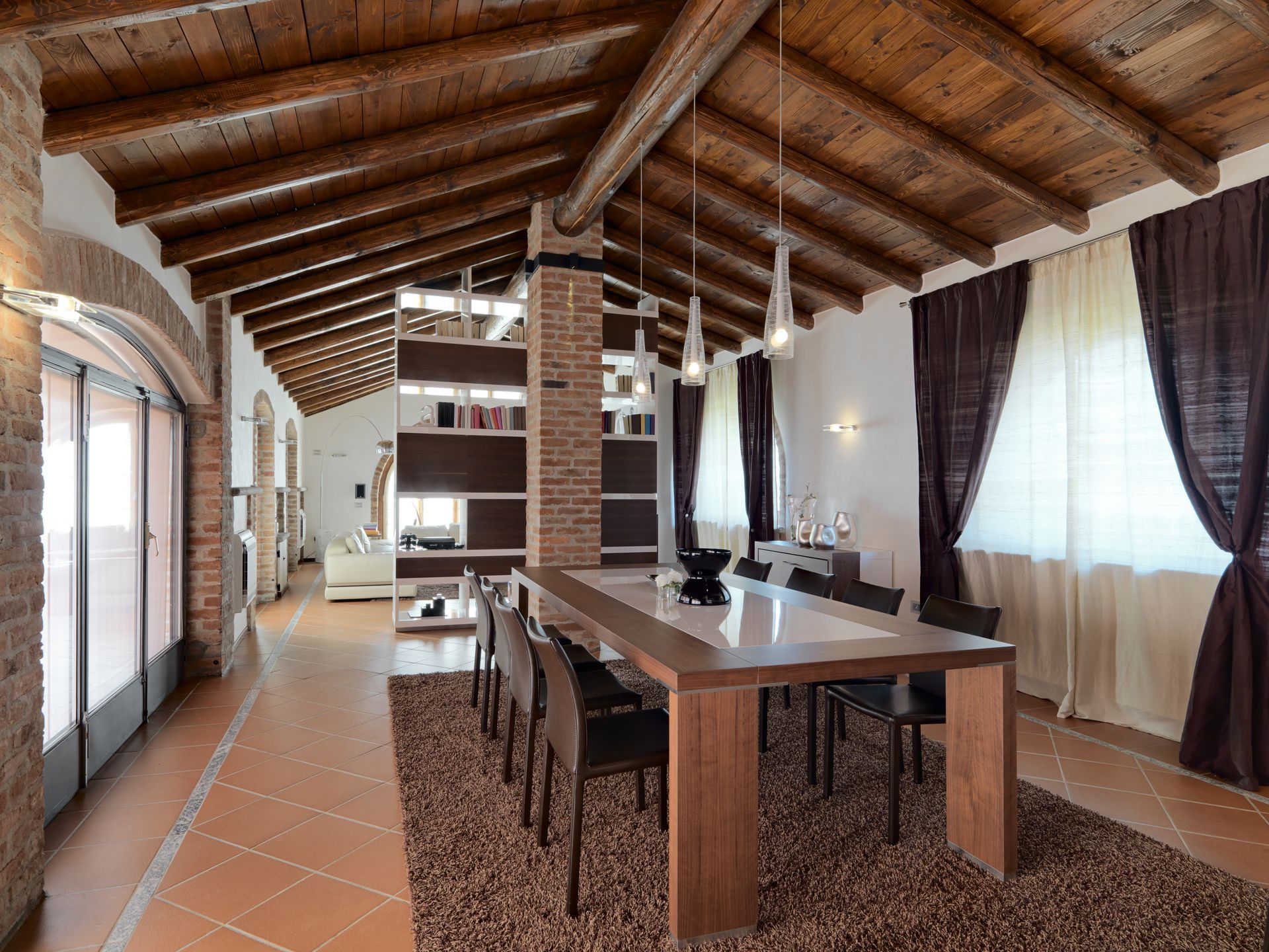 Rustic dining room with long wooden table, terracotta tile floor, exposed beams, and large curtained windows
