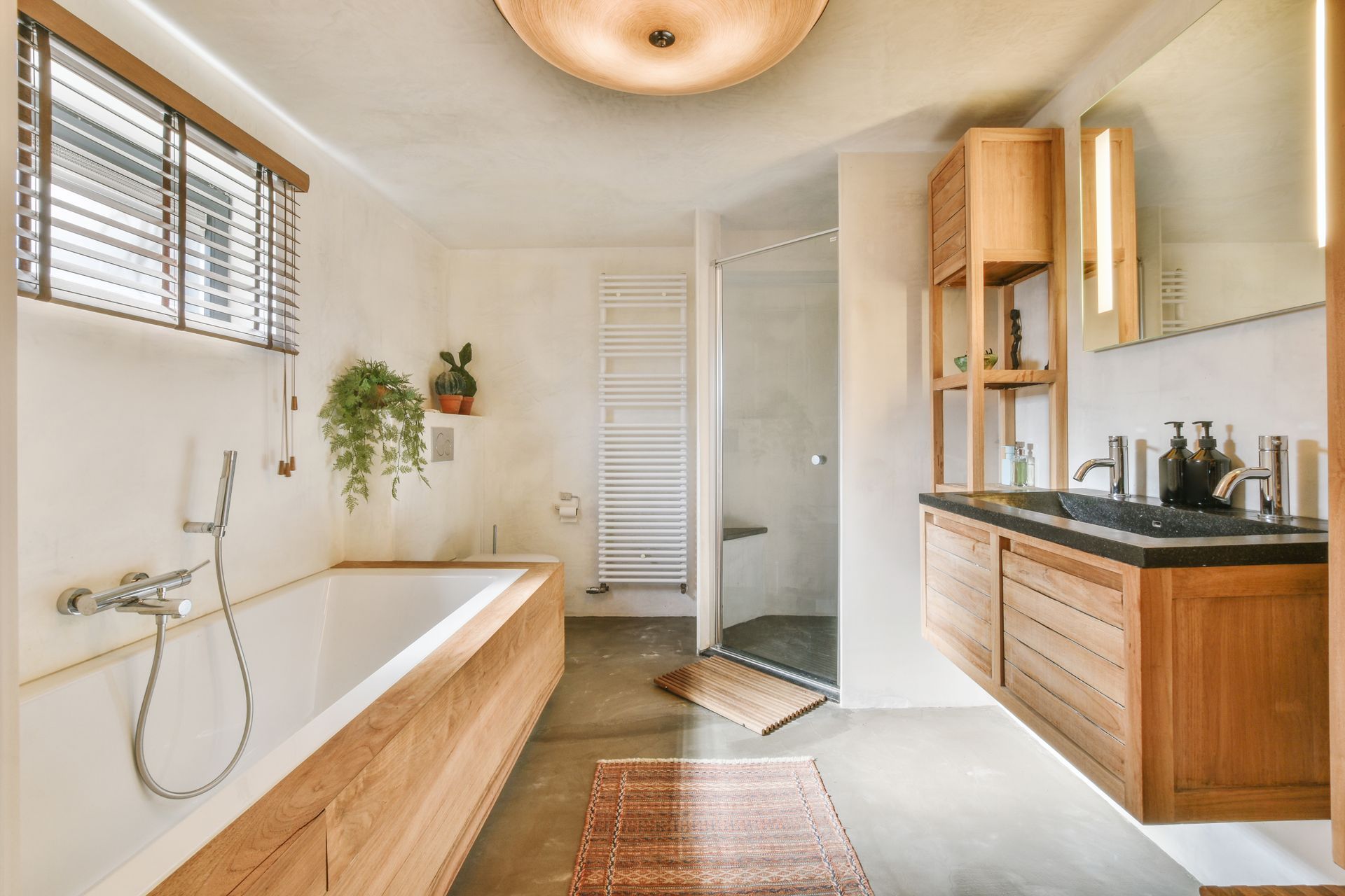 Bright modern bathroom with a freestanding tub, wood vanity, large mirror, and window blinds.