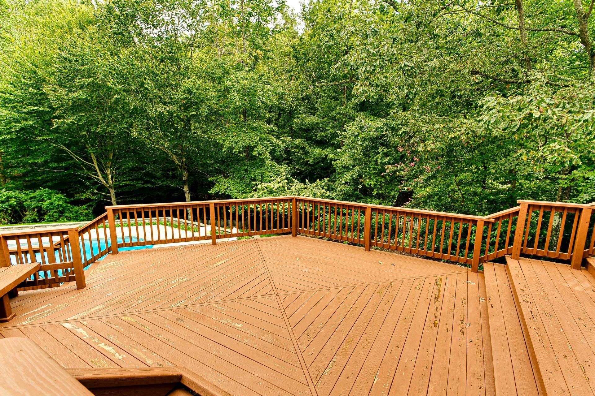 Wooden deck with railings overlooking a lush green forest and a small pool on the left