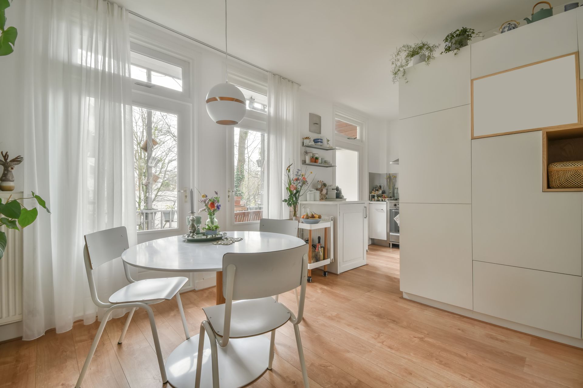 Bright minimalist dining room with white chairs, round table, wooden floor, and large windows with curtains