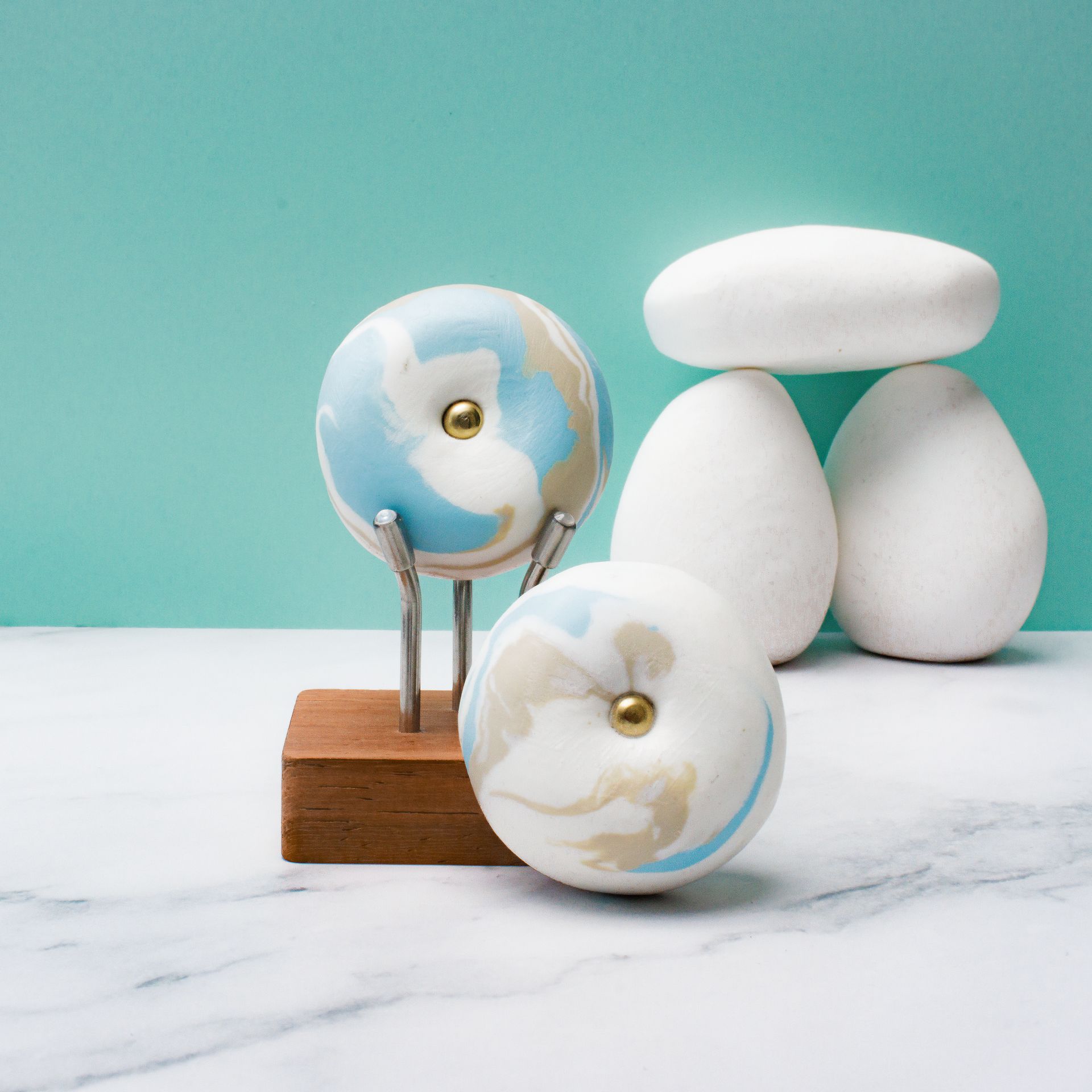 Witte abstracte kiezels en twee ronde glazen ornamenten op een houten blok tegen een turquoise achtergrond.