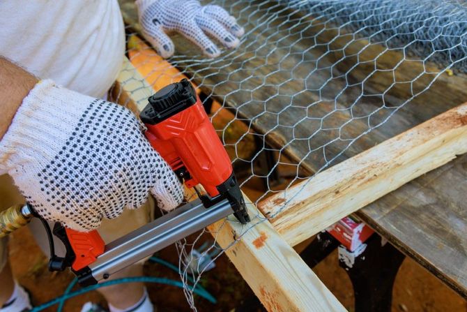 Person using a nail gun to attach chicken wire to wooden frame, wearing gloves.