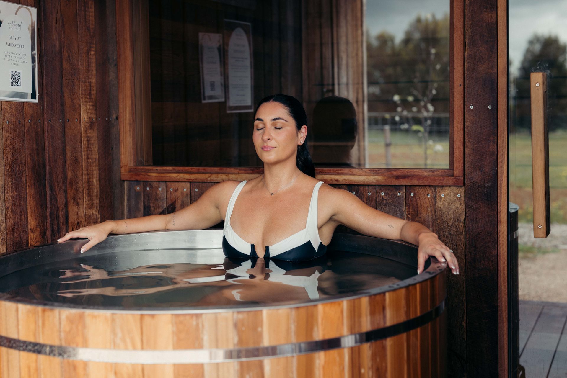 Woman in a swimsuit relaxing in a wooden hot tub, eyes closed, arms outstretched, with a natural setting in background.