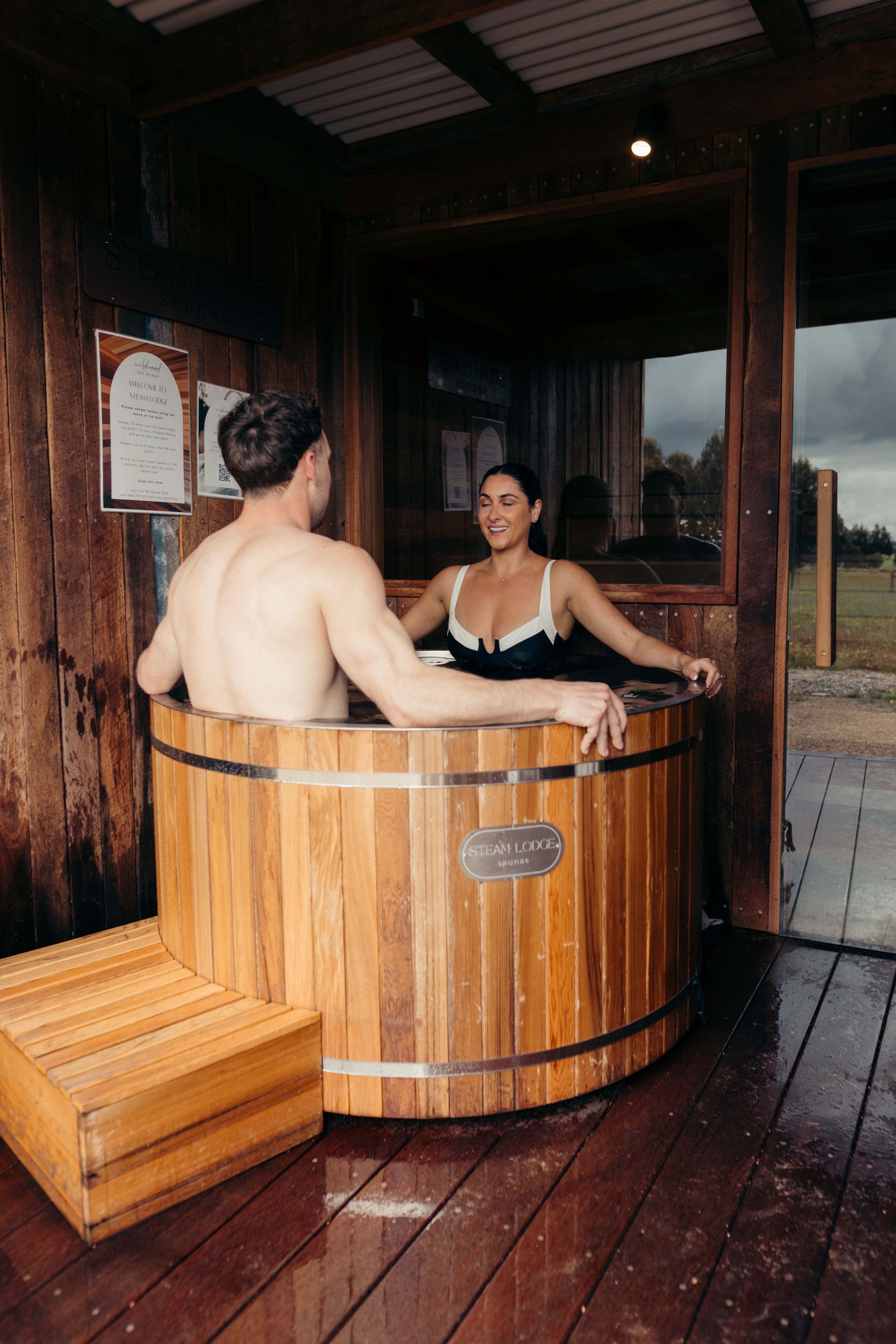 Couple in a wooden hot tub, smiling, in a rustic outdoor setting.