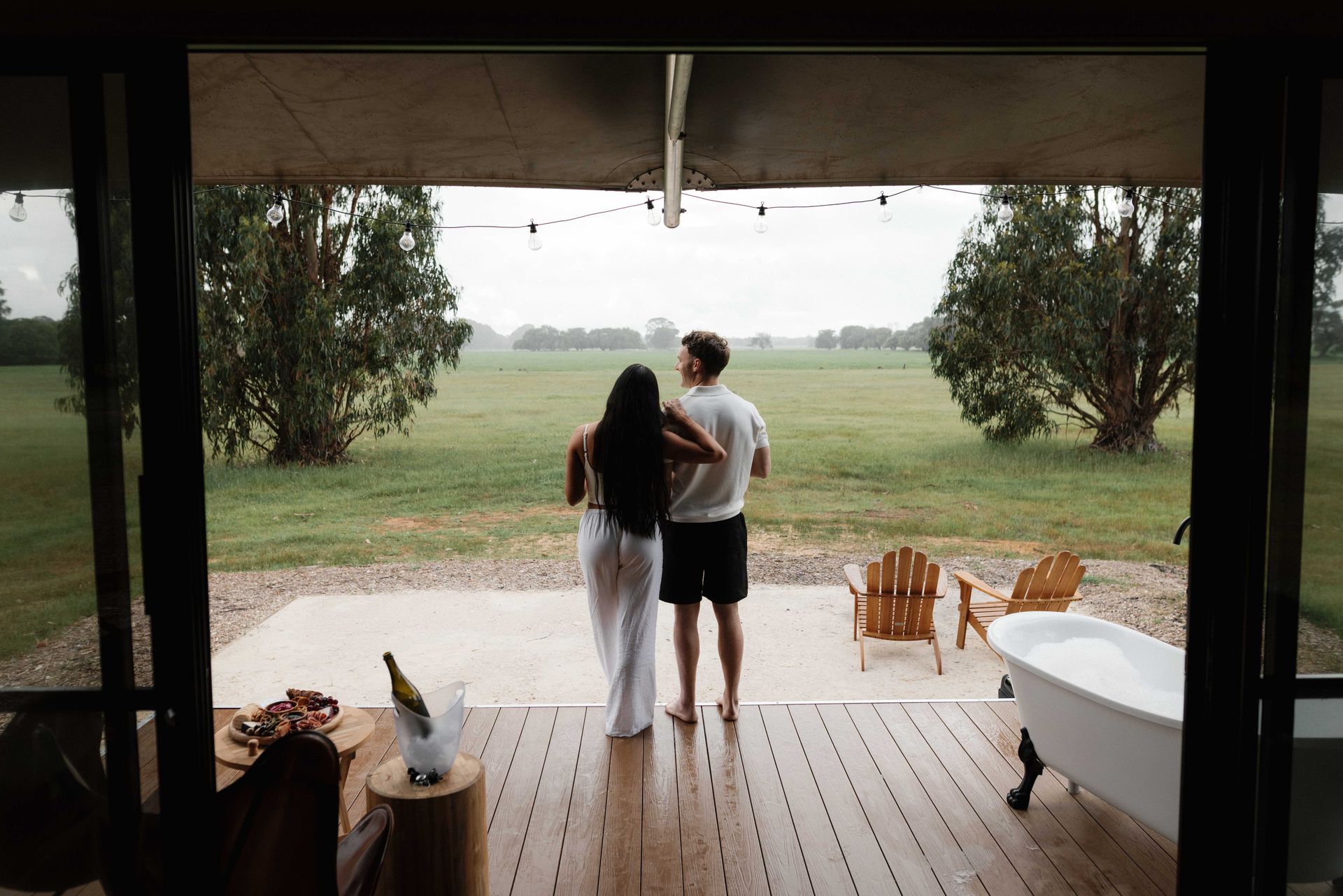 Couple standing on a porch looking at a rainy field. Wine and a bathtub on the porch.