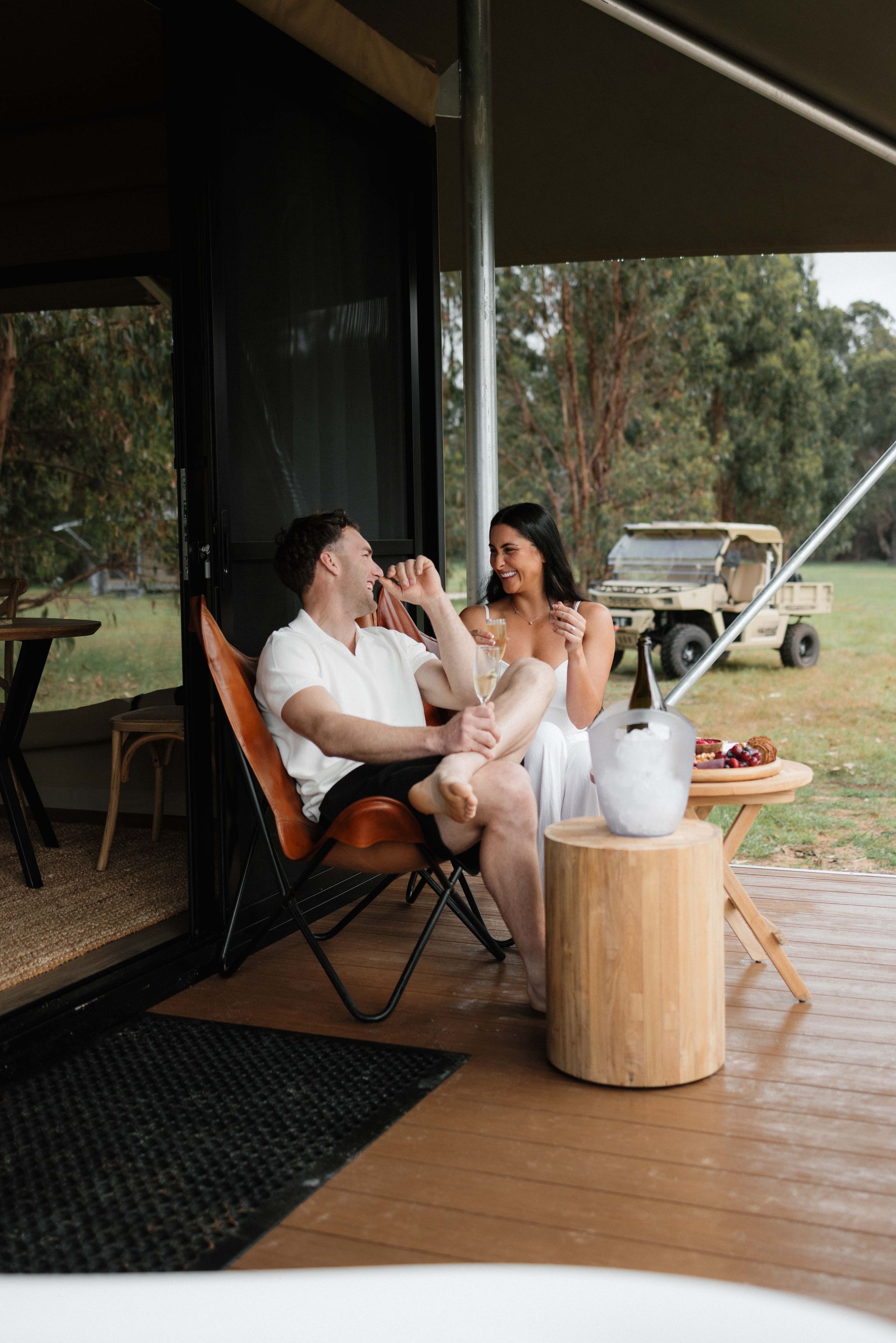 Couple seated outdoors, drinking, near a small table with champagne and grapes.