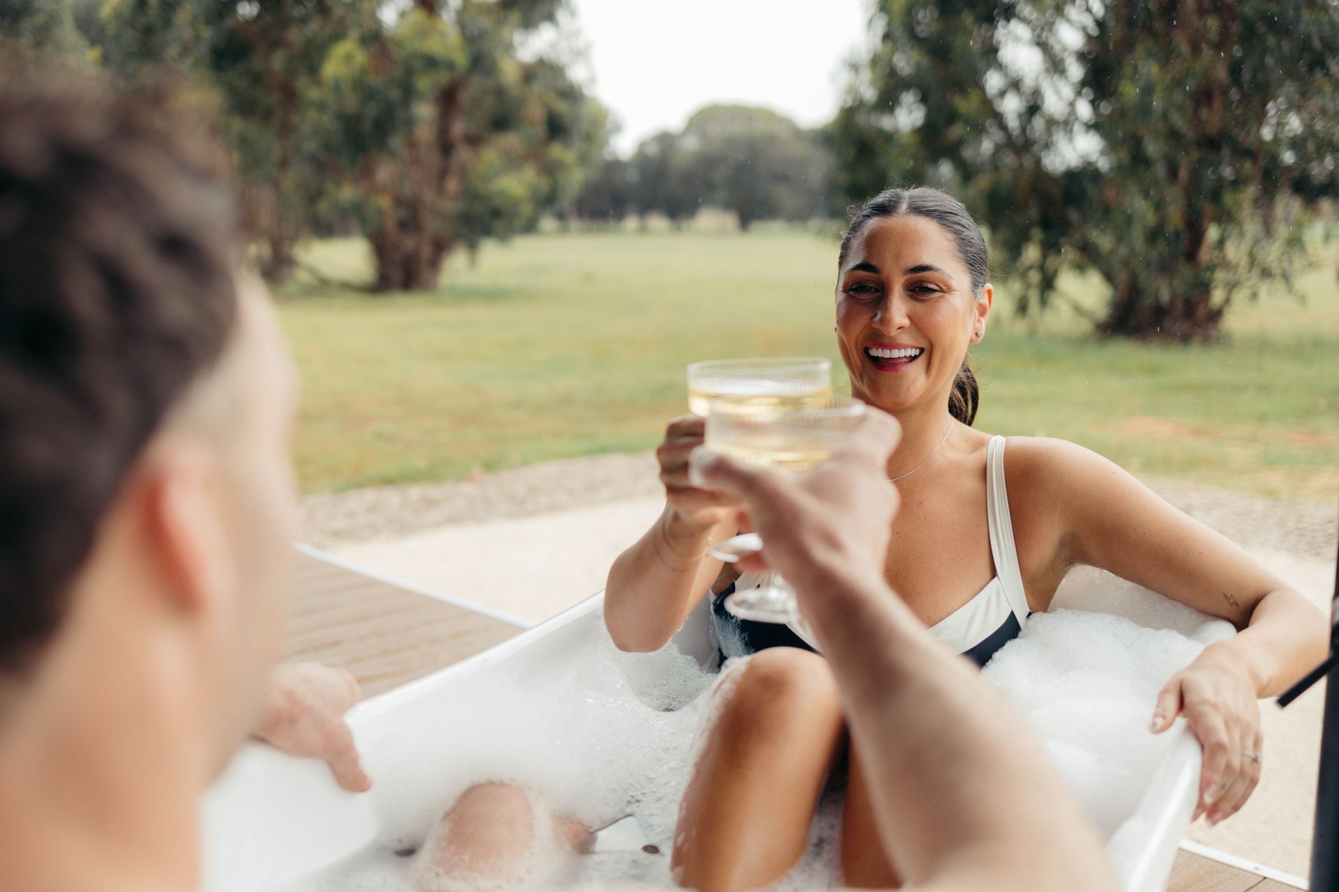 Woman in a bathtub cheers with a glass of wine, smiling at someone out of frame. Outdoor setting.