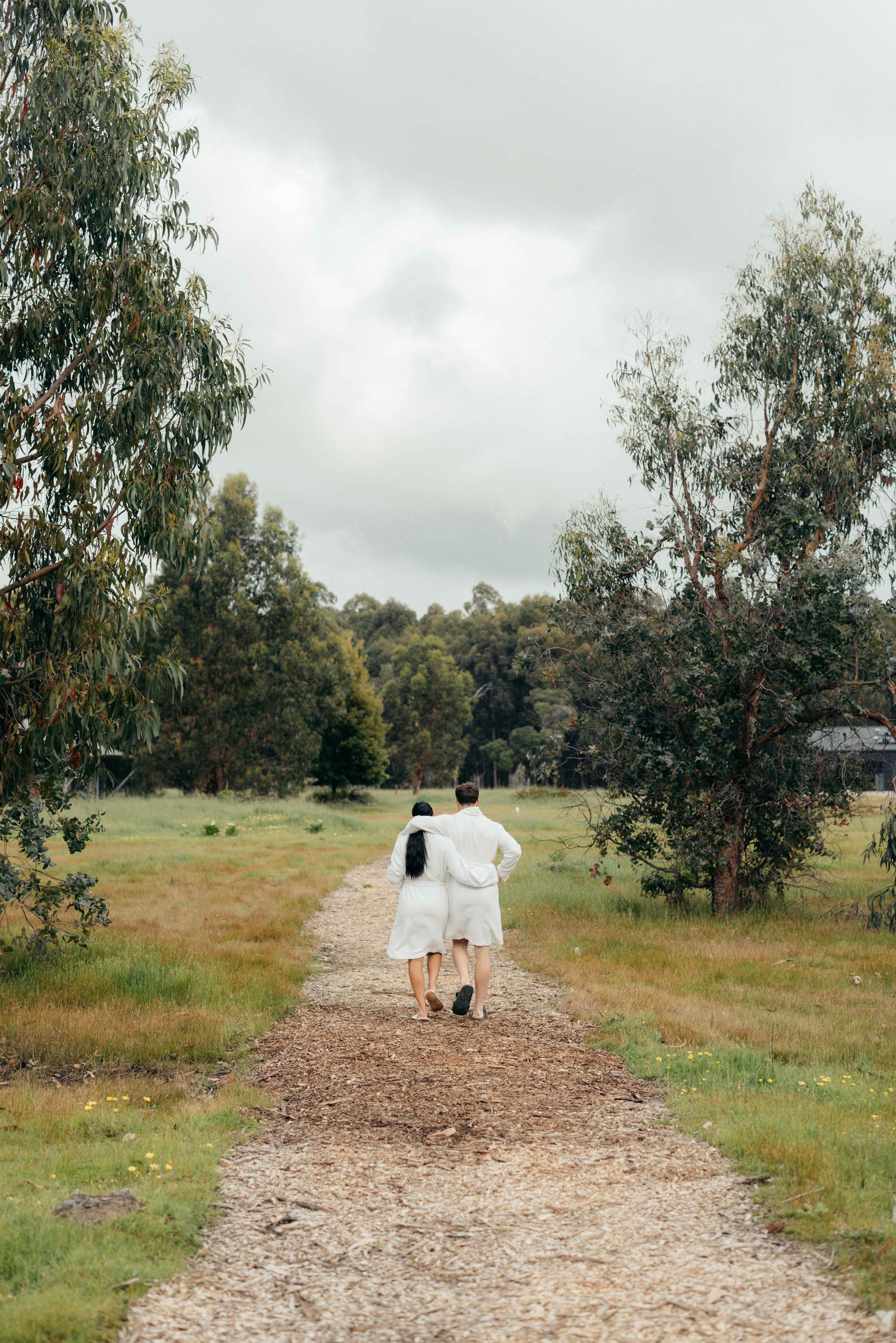 Two people walking on a gravel path through a field, trees on either side, overcast sky.