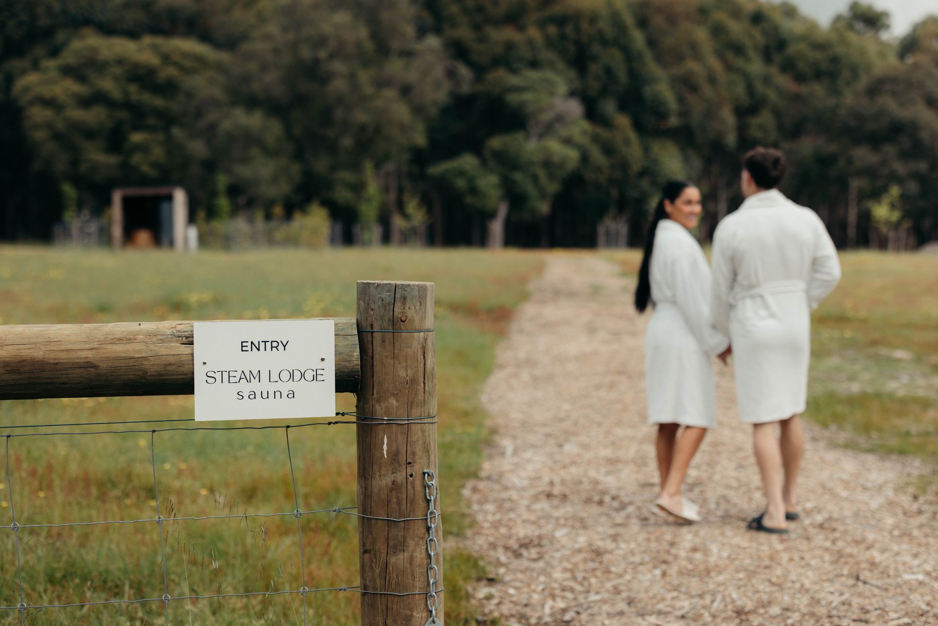 Couple in white robes walking on a gravel path, holding hands, with a sign on a wooden fence.