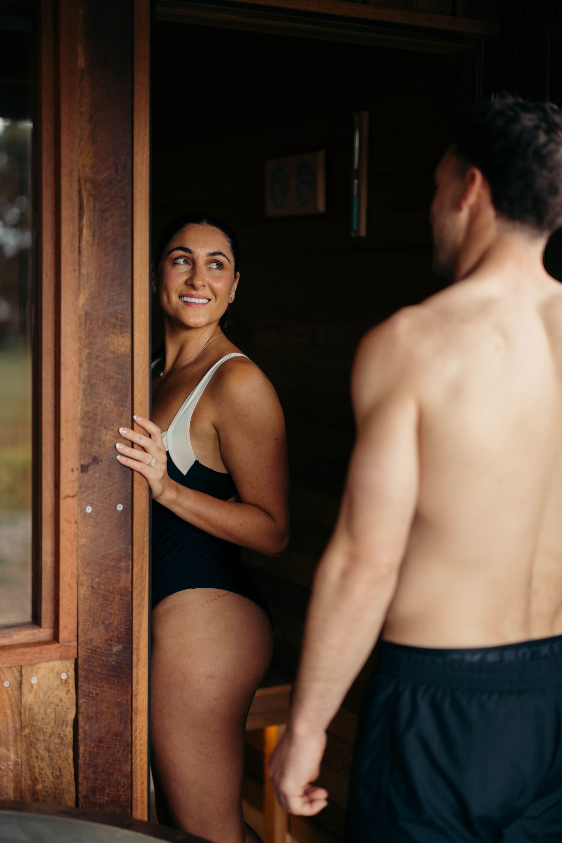 Woman in swimsuit smiles at man, standing in sauna doorway.