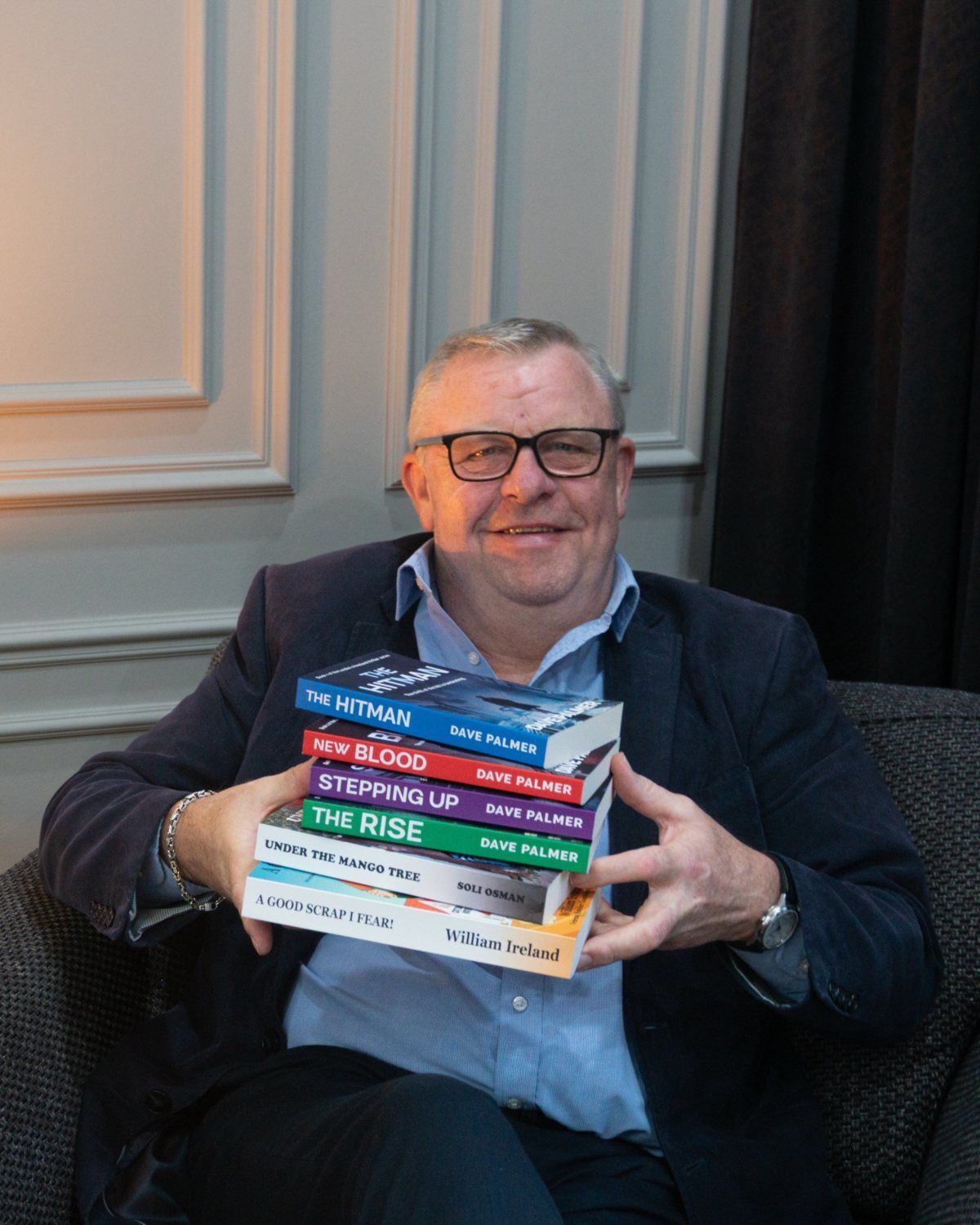 Dave Palmer smiling in an armchair, holding a stack of books: his own novels and two of his clients' books