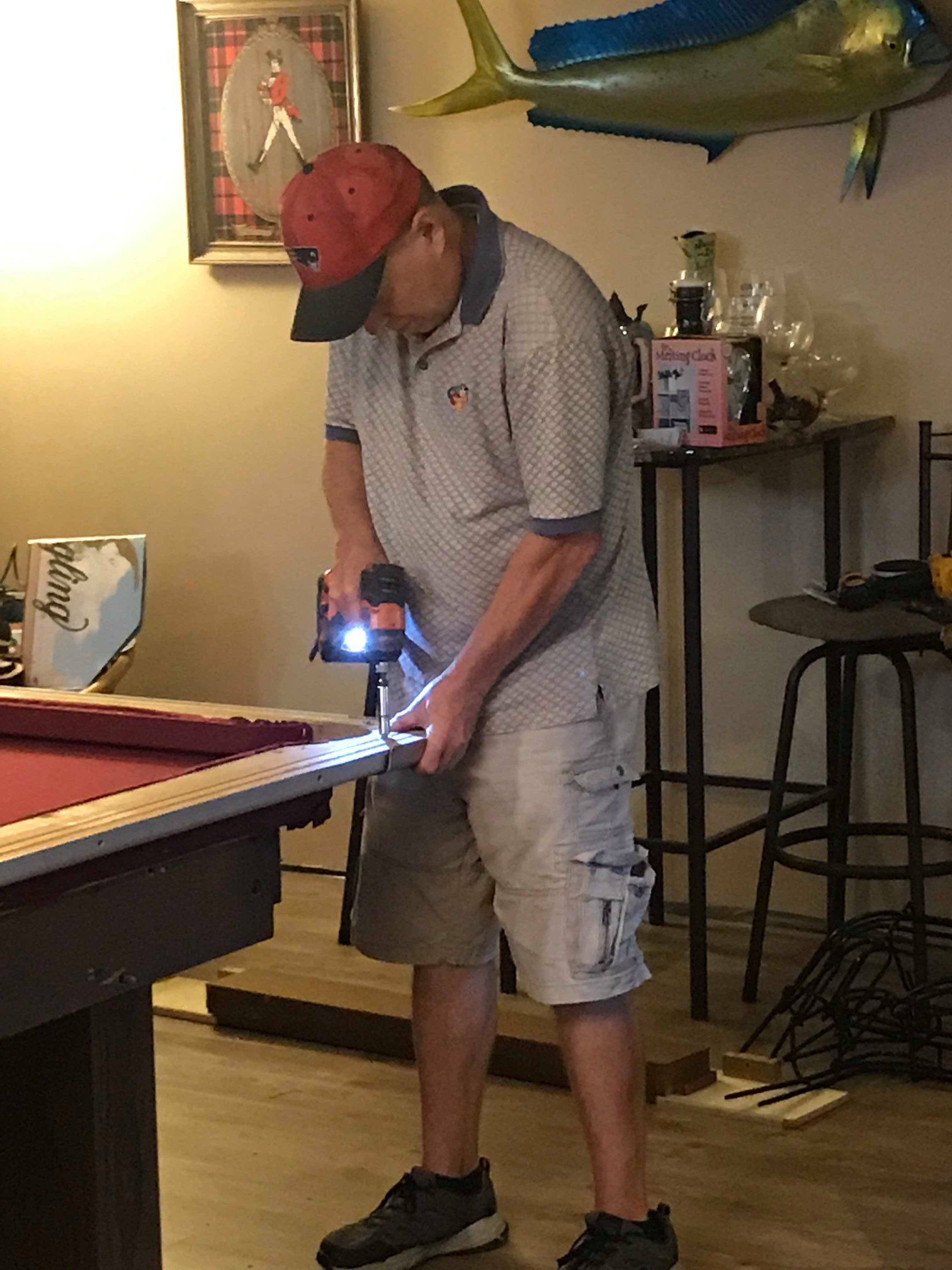 Man in red cap using a power drill on a pool table in a room.