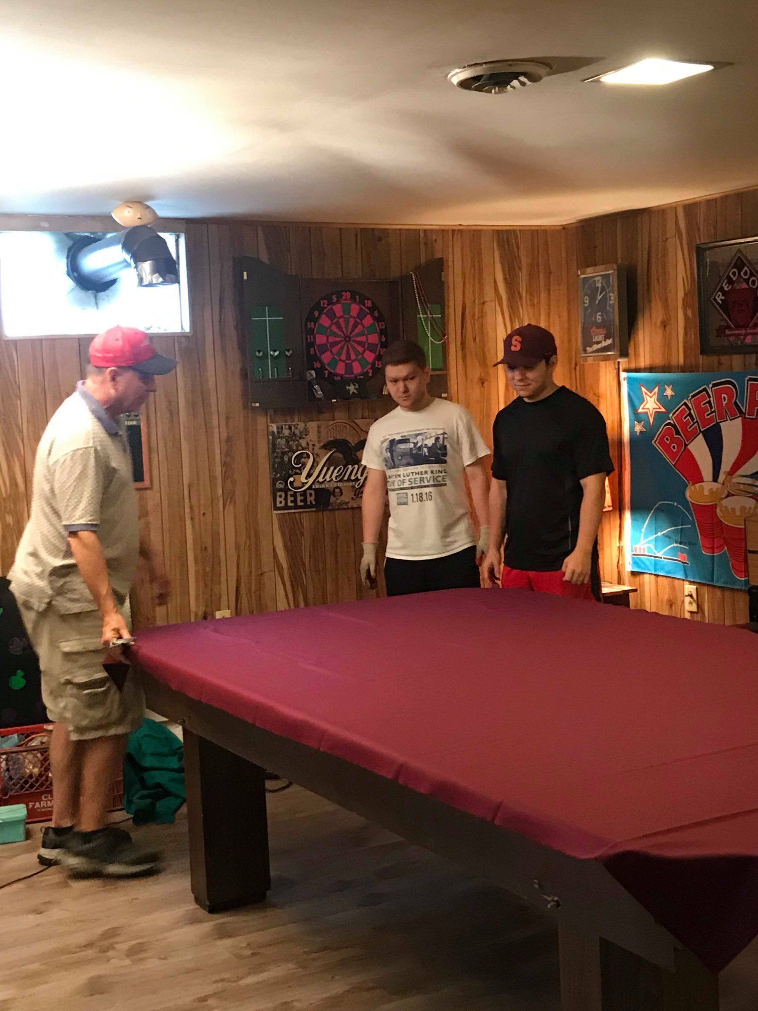 Three people covering a maroon pool table in a wood-paneled basement. Three people covering a maroon pool table in a wood-paneled basement.