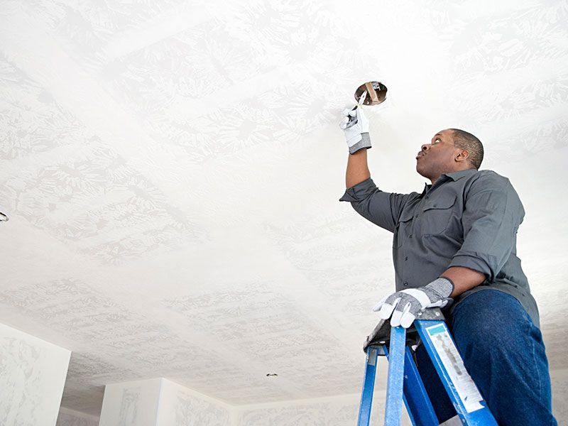 Person on a ladder working on a ceiling electrical fixture. Wearing gloves, gray shirt, and jeans.