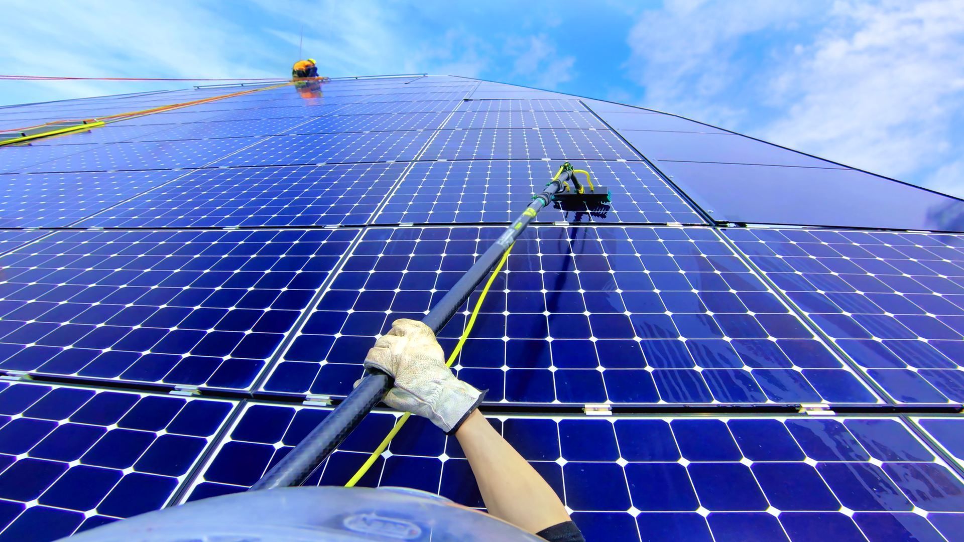 Person cleaning solar panels with a long-handled brush on a rooftop under a blue sky.