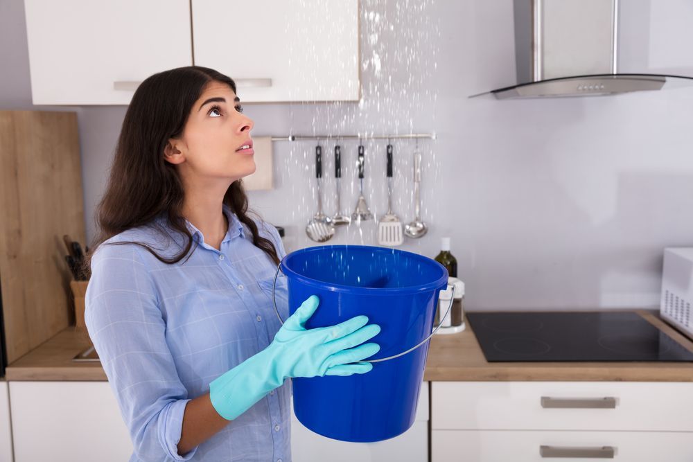 A Woman is Holding a Blue Bucket With Water Coming Out of the Ceiling — Accredited Plumbing in Mollymook, NSW