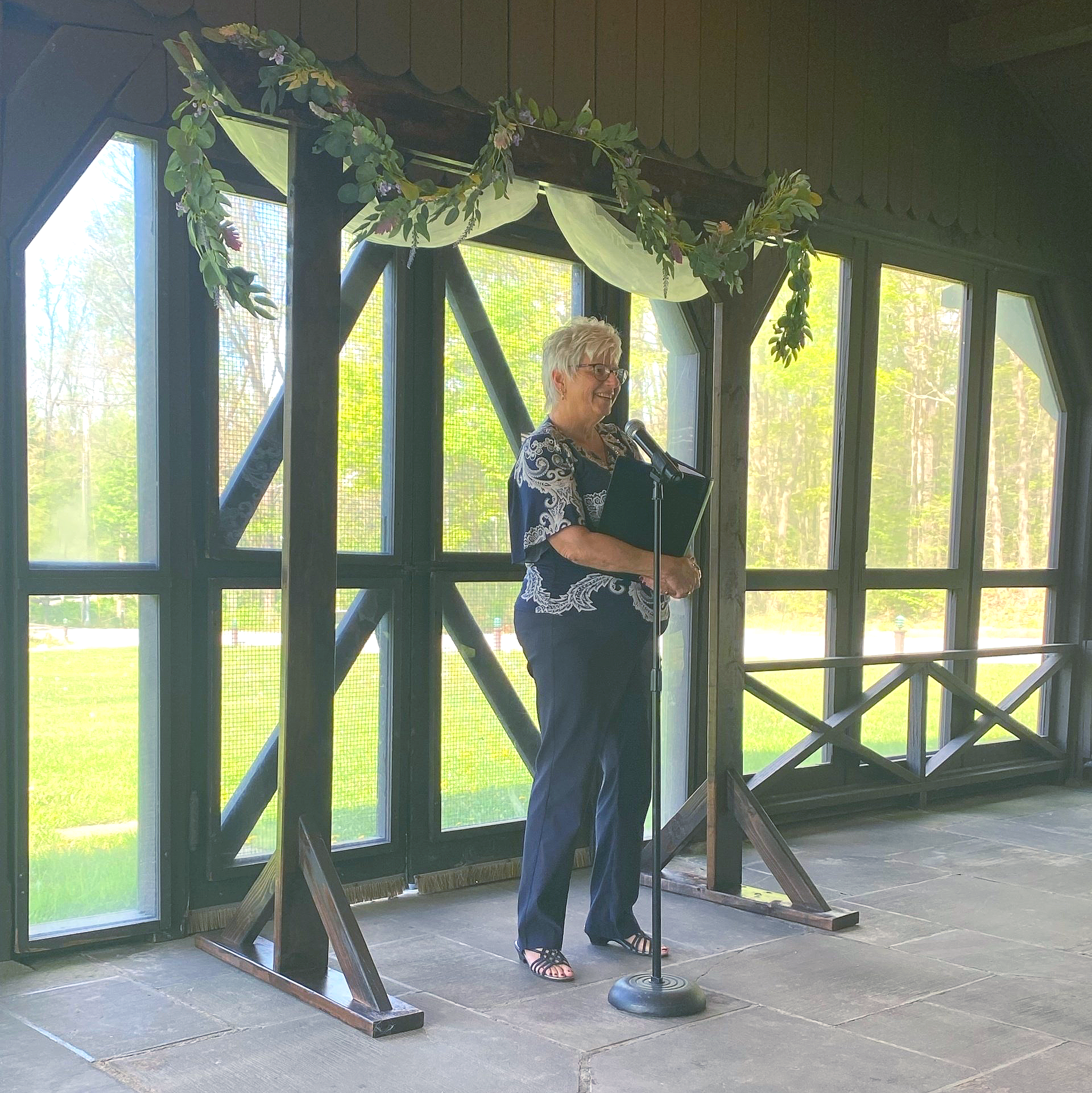 Woman stands at microphone beneath a decorated wooden archway, inside a room with large windows.