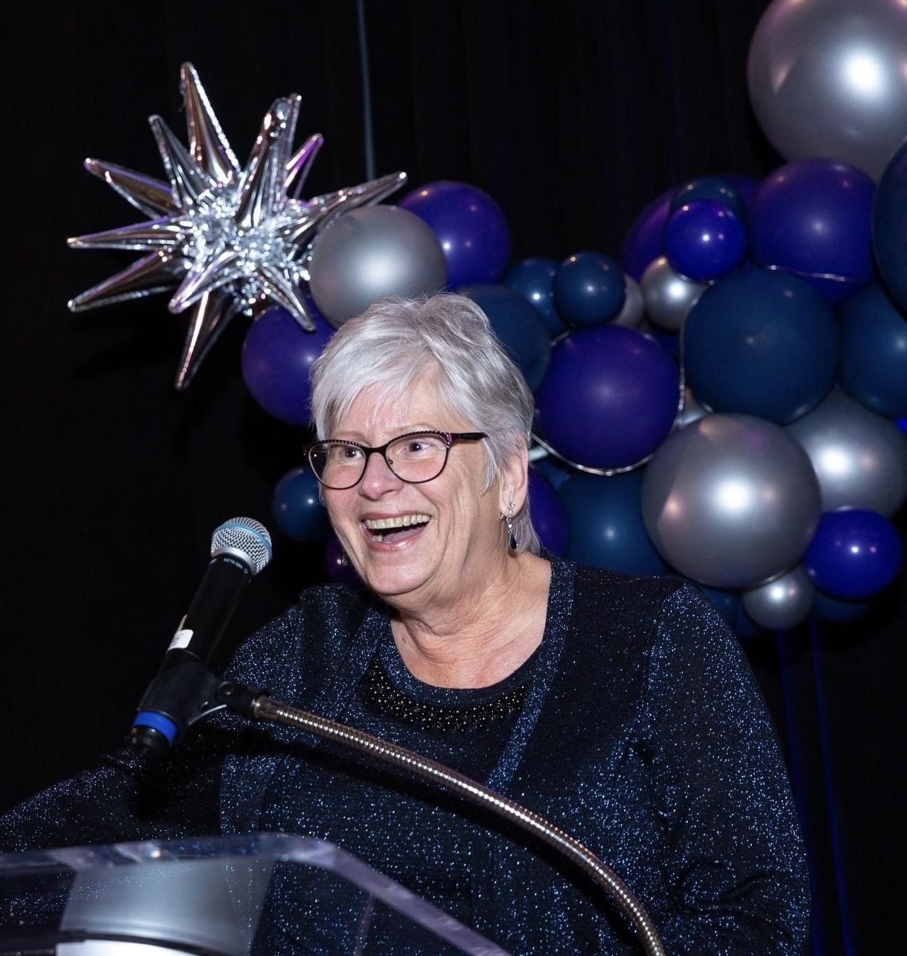 Woman speaking at a podium, smiling, with a balloon arch in the background.