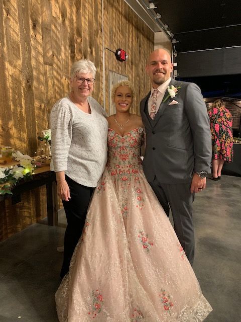 Woman in pink dress with floral embroidery poses with a man in gray suit and another woman indoors.