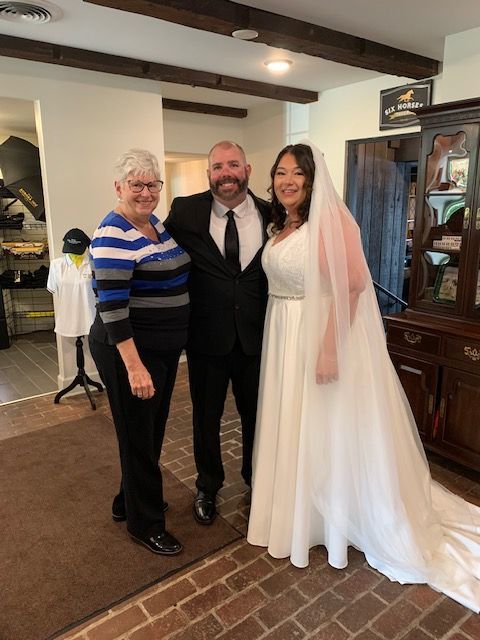 A bride, groom, and woman pose indoors. The bride wears a white dress and veil, the groom a black suit.