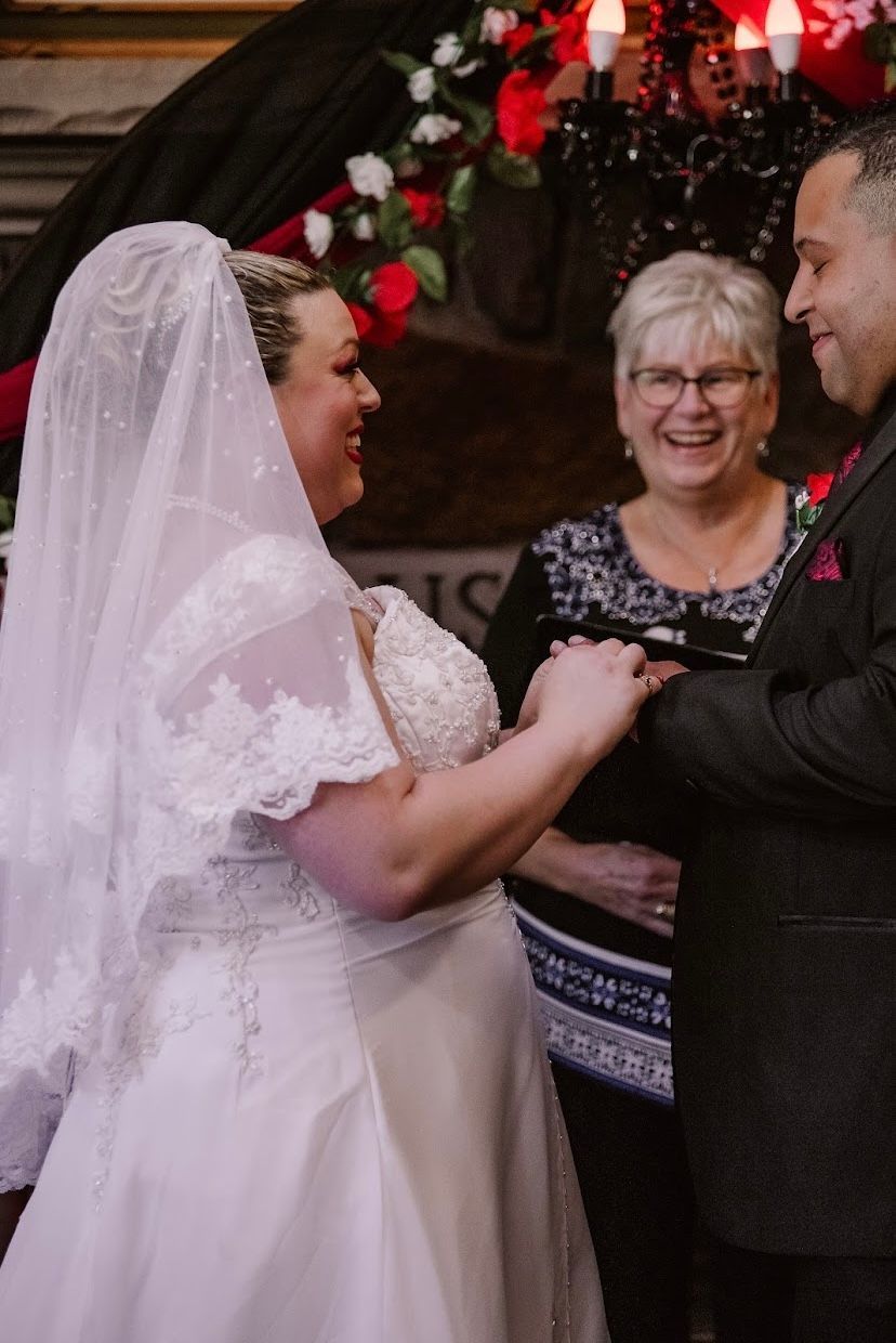 Bride and groom exchange rings during wedding ceremony; officiant smiles.