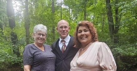 Three people pose outdoors in front of trees. Man in suit, two women in dresses, smiling.