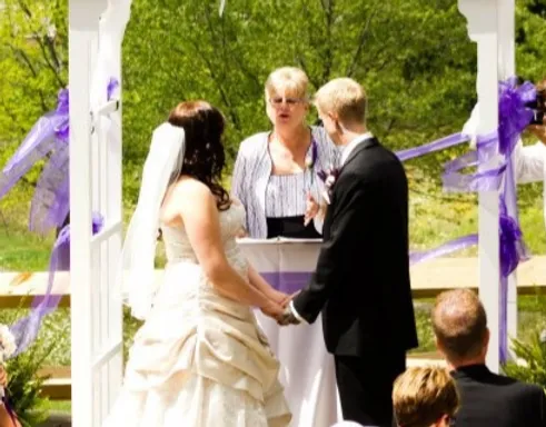 Couple holding hands during outdoor wedding ceremony; officiant stands before them under an arbor.