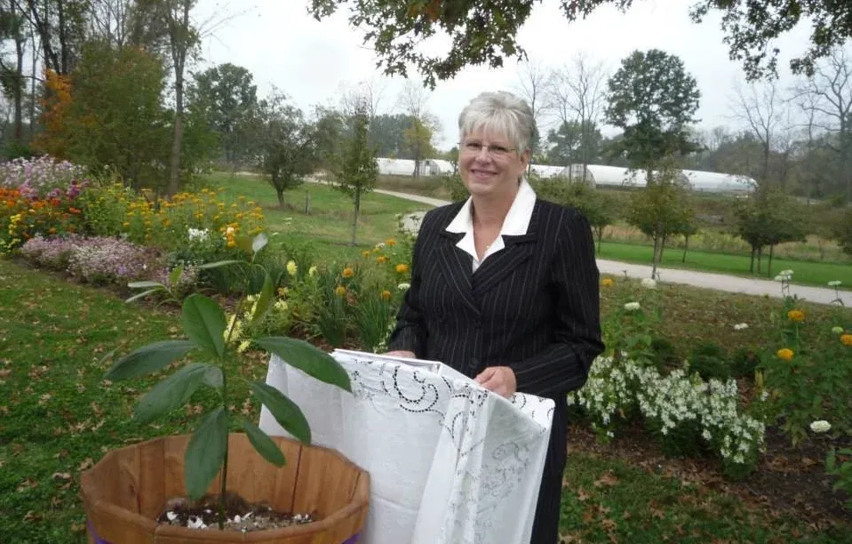 Woman in a suit stands in a garden holding a white cloth; a potted sapling is in front.
