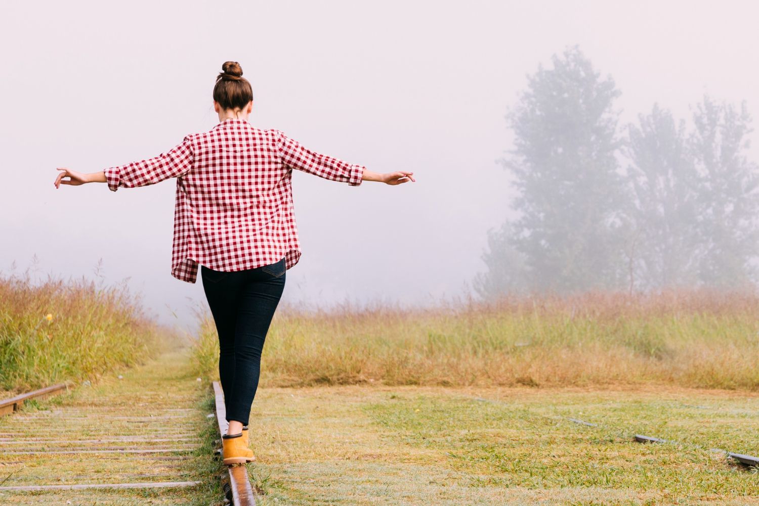 Person in a red plaid shirt balances on a narrow path in a foggy field with arms outstretched.