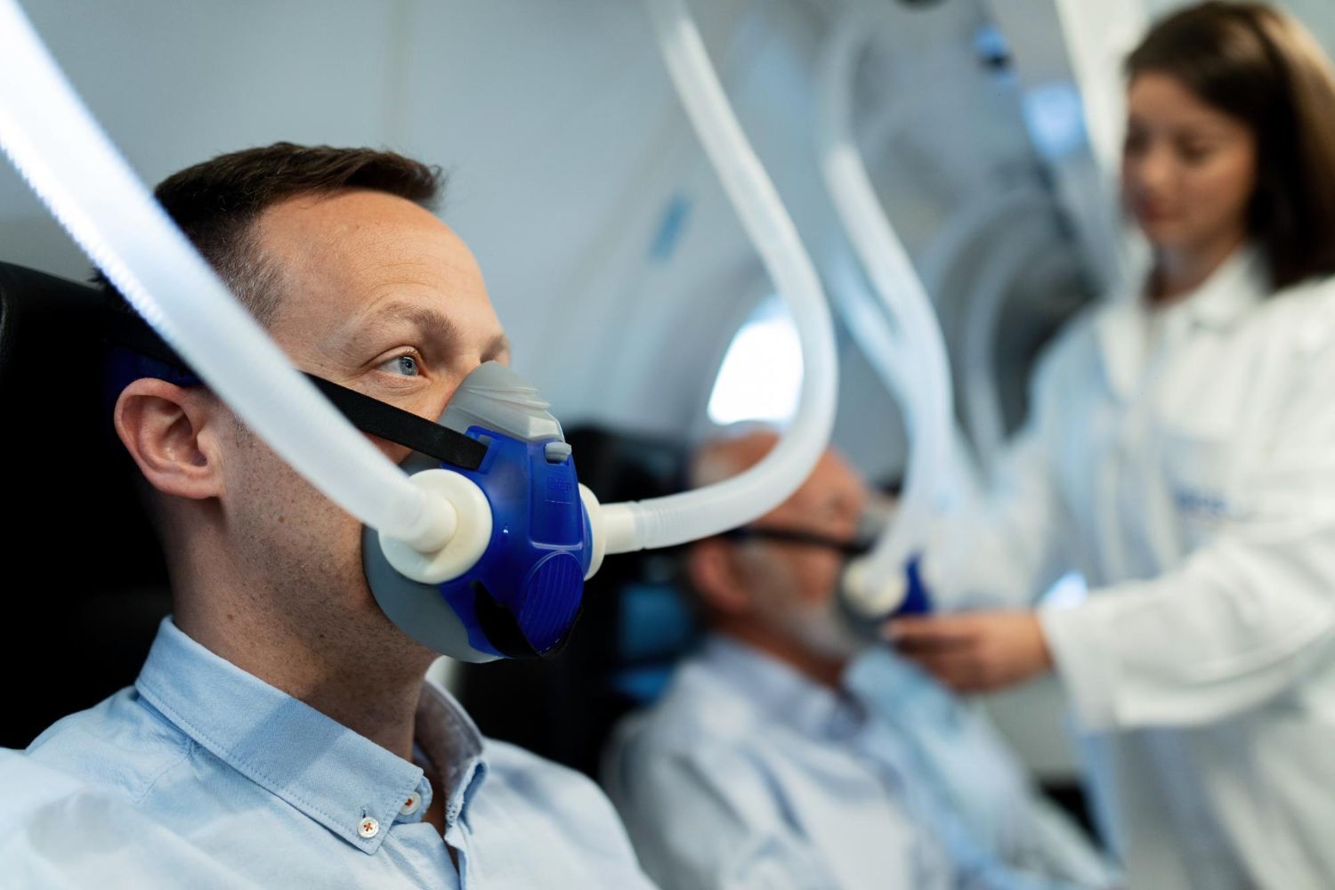 Patient wearing blue oxygen mask in hospital while medical staff monitor equipment in the background